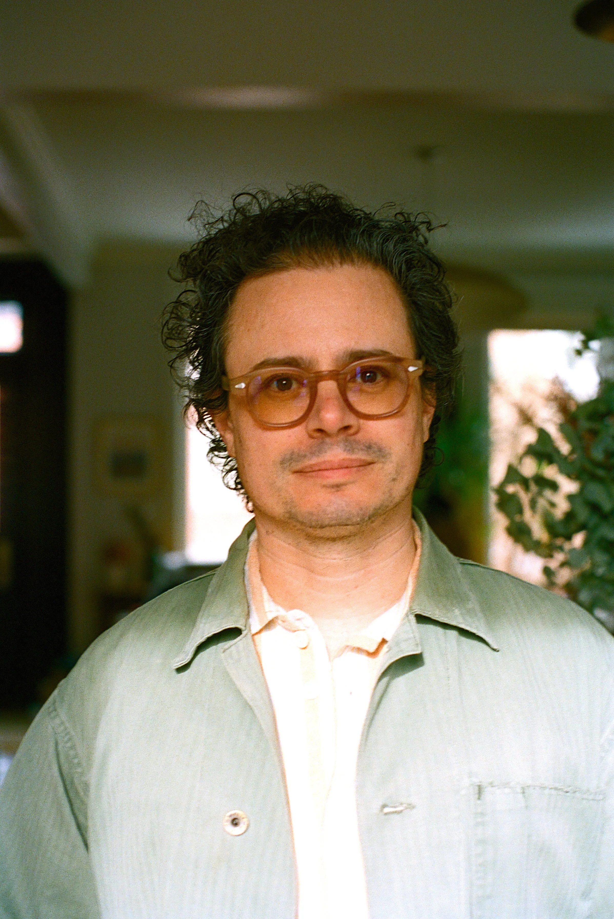 A man with curly dark hair wearing glasses with a beige frame, a light green jacket, and a white shirt, standing indoors.