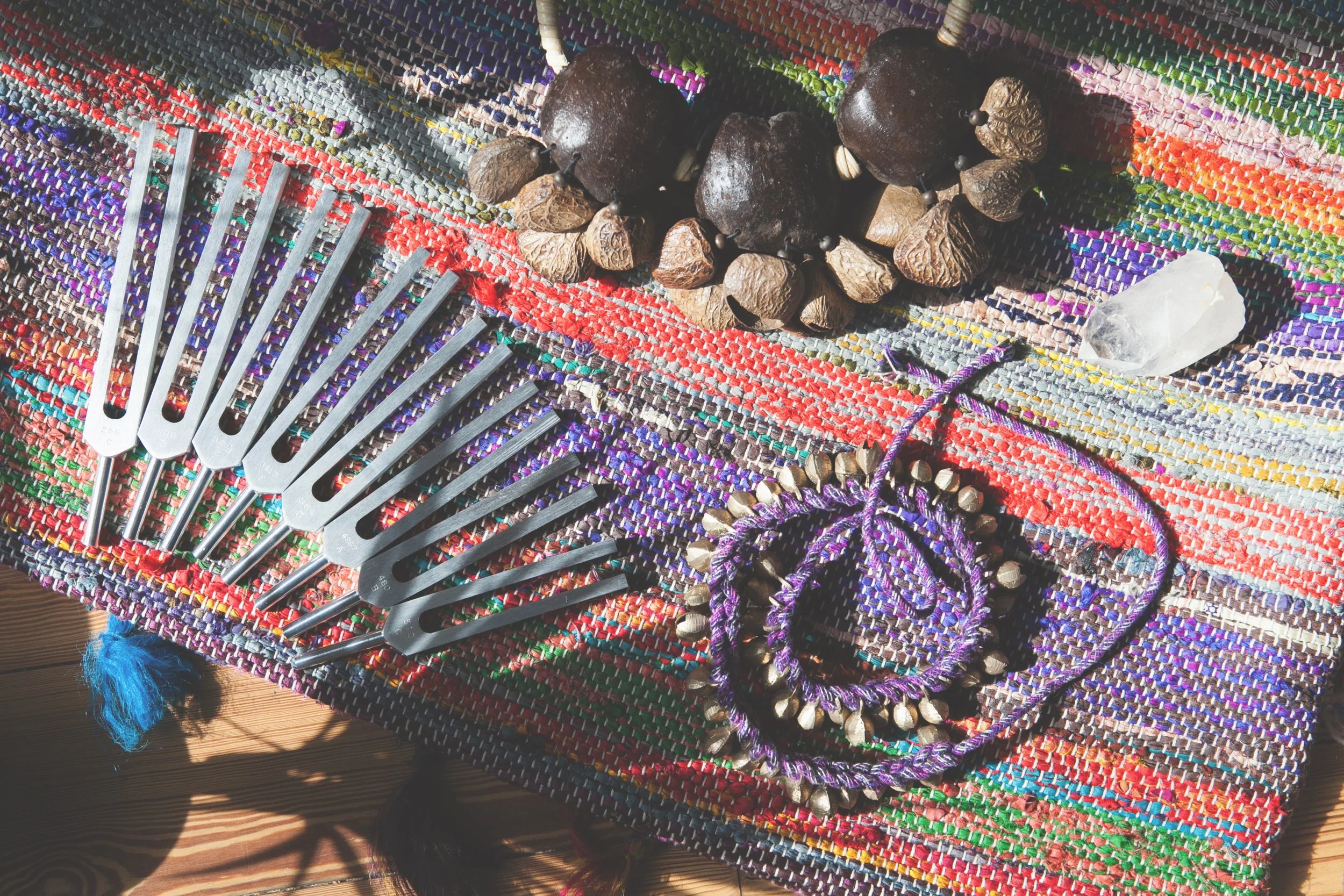 Colorful woven fabric with a set of metal forks, a cluster of dried seed pods, a clear quartz crystal, and a purple drawstring with small metallic beads, all arranged on it.