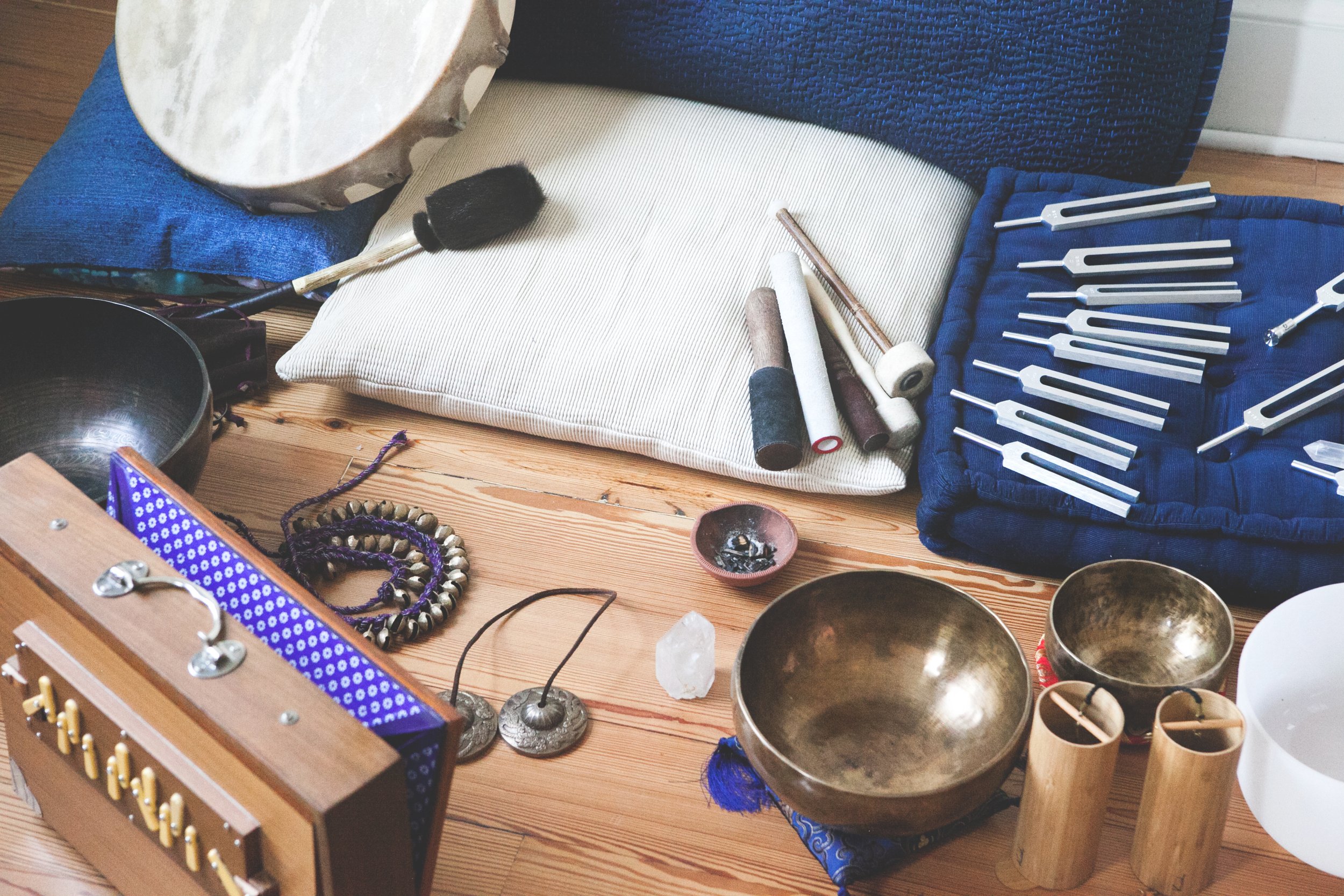 Collection of musical instruments and accessories on a wooden floor, including singing bowls, drums, a mallet, a drumstick, and a wooden chime set, with some pillows and fabric.