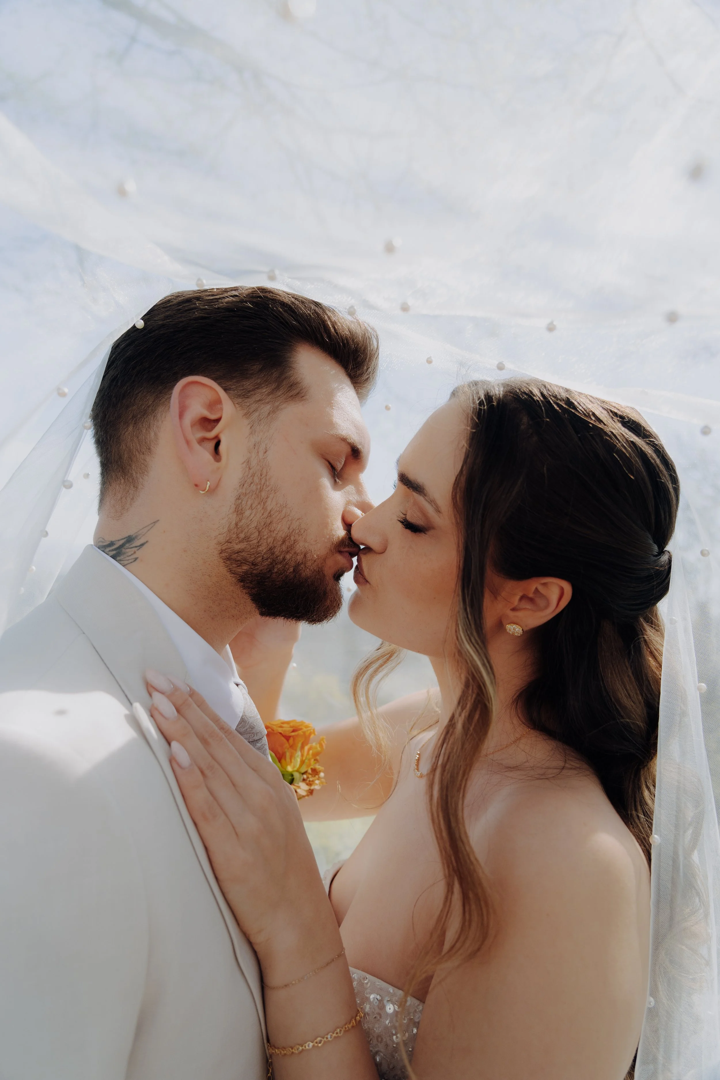 Ein Paar küsst sich bei einer Hochzeit im Freien, unter einem weißen Tüll mit Perlen, im Hintergrund sind Bäume sichtbar.
