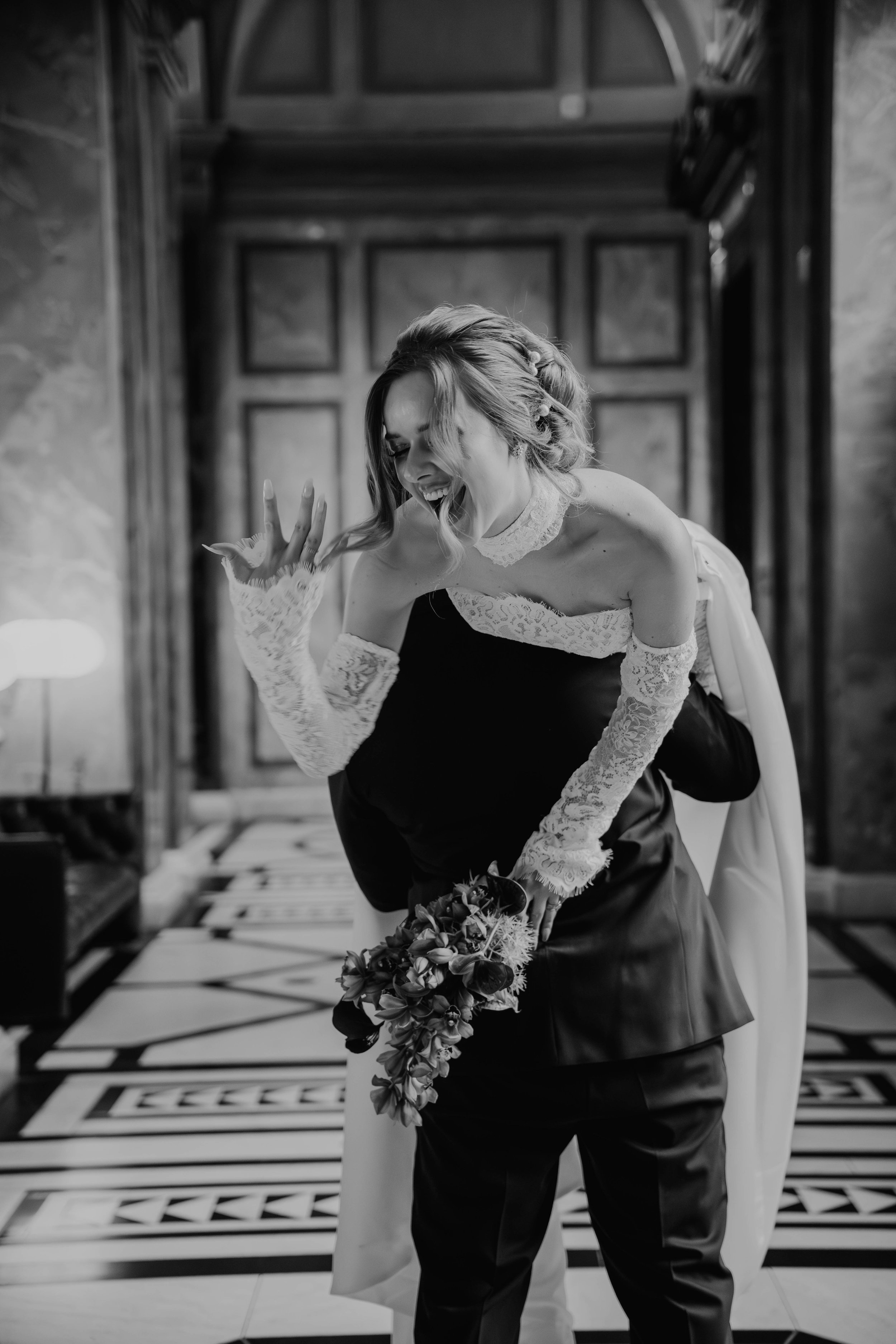Frau in Brautkleid sitzt auf einem Mann mit Smoking, hält einen Blumenstrauß, lächelt und lacht, im Hintergrund ein Holz-Türen in einem Raum.