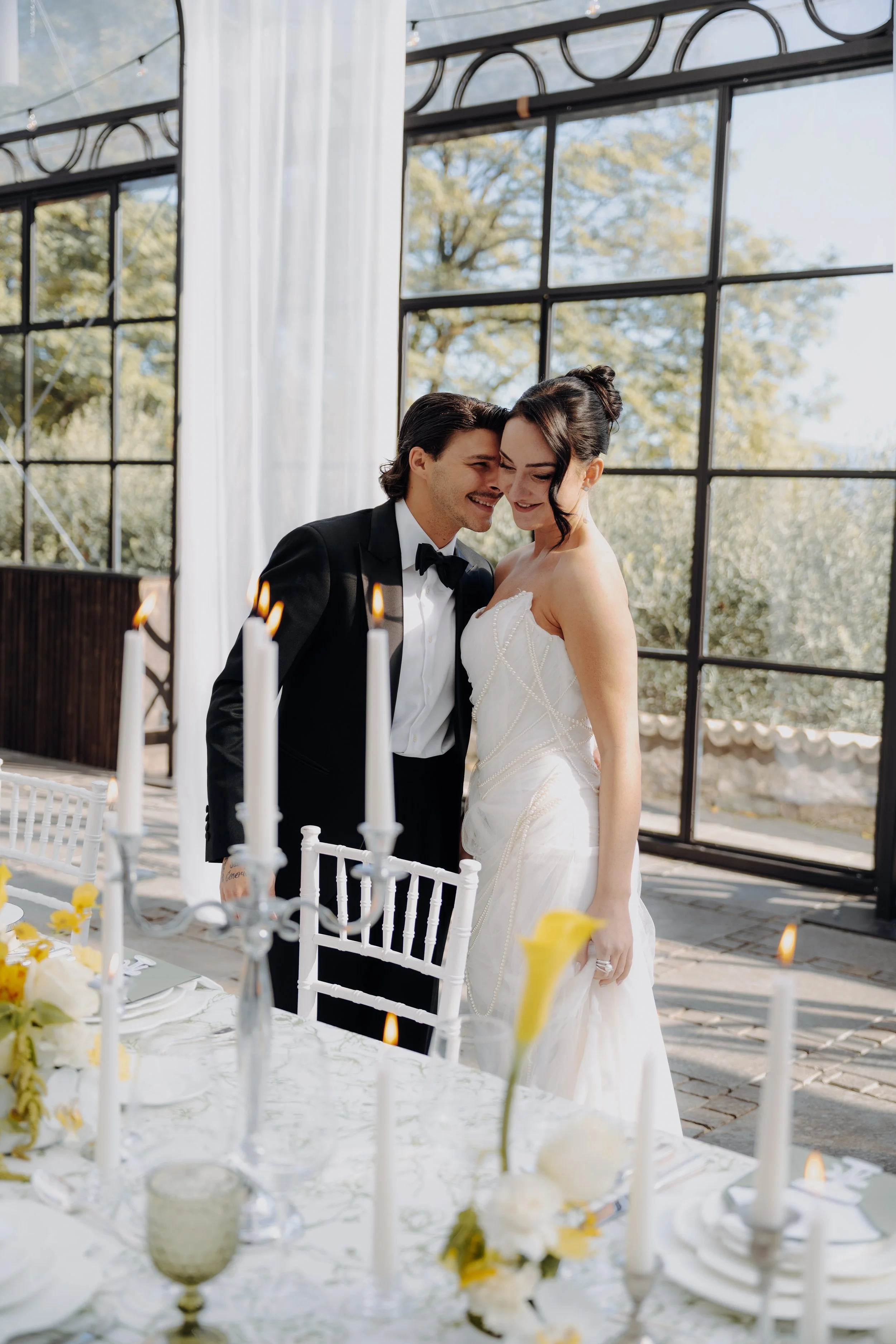 Braut und Bräutigam bei ihrer Hochzeit, die den Kopf aneinander legen, in einem Raum mit großen Fenstern und Blick auf Bäume, bei Sonnenlicht, auf einer weißen Hochzeitstafel mit Kerzen und Blumen. 