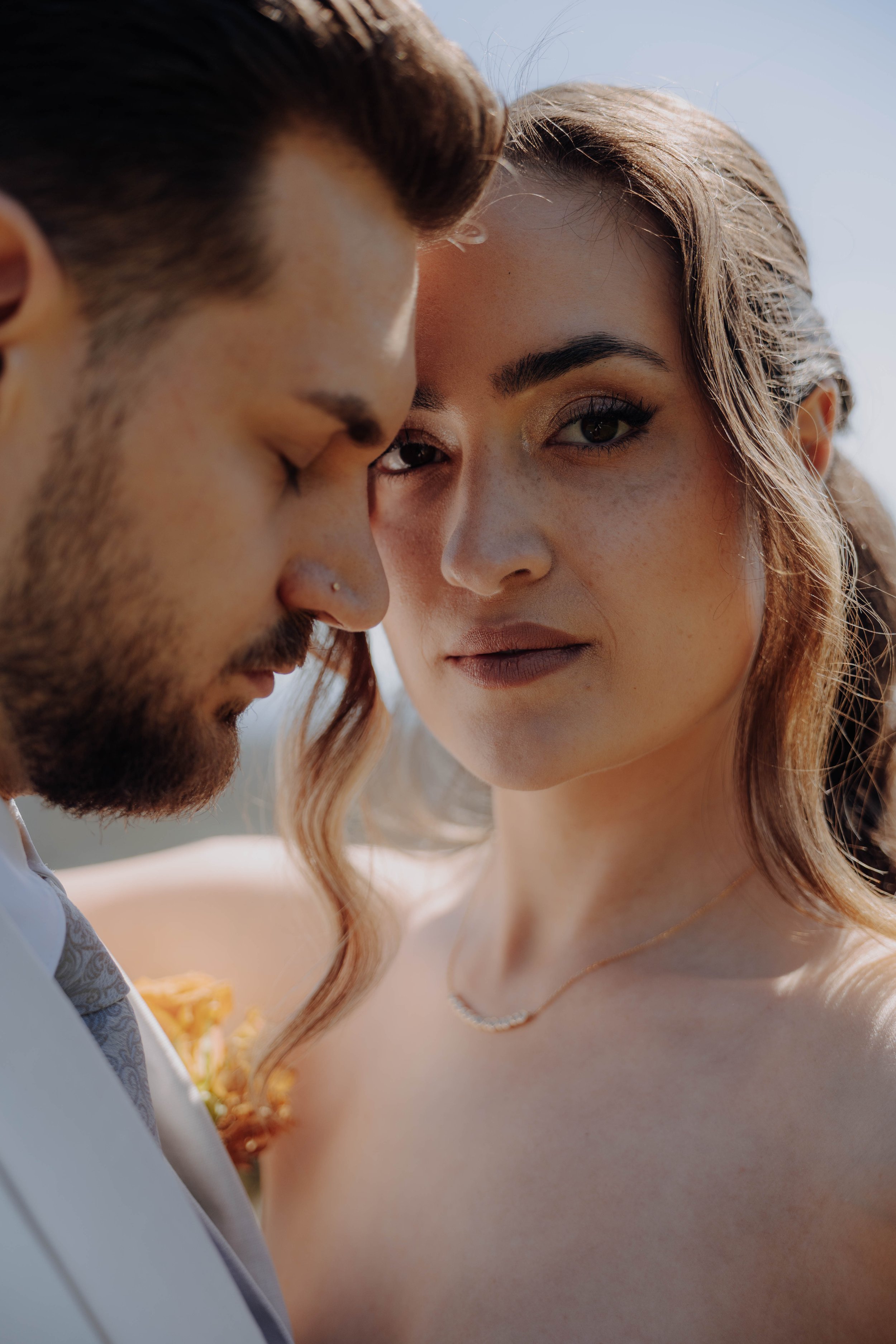 Ein Paar steht sich nahe bei Sonnenlicht, der Mann mit dunklem Haar und Bart, die Frau mit lockigem, hellbraunem Haar und leichter Make-up, beide schauen intensiv. Der Hintergrund ist unscharf, Himmel sichtbar.