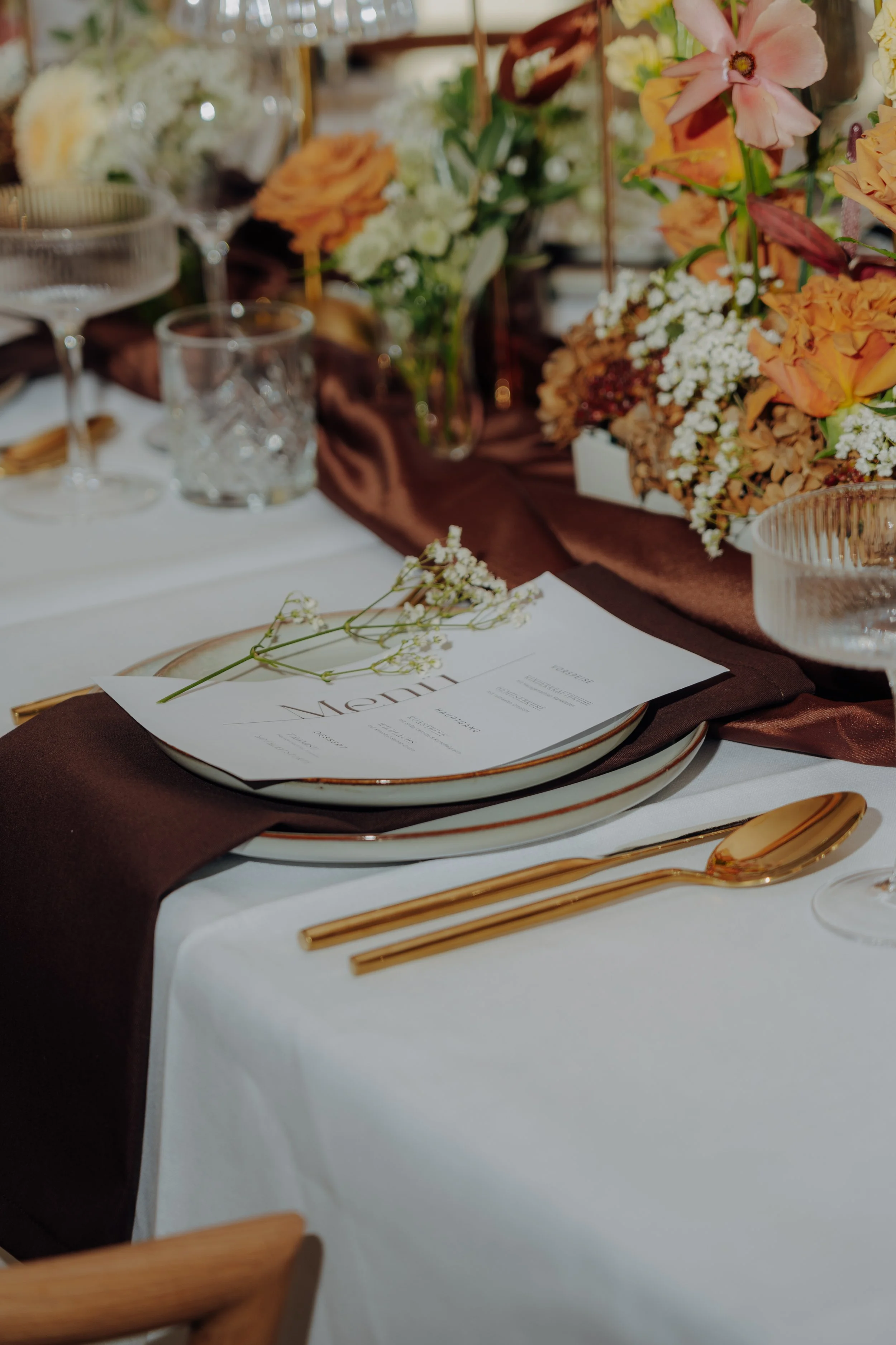 Tisch mit weißen Tellern, goldene Besteck, braune Stoffserviette, Blumenarrangement und Menükarte mit kleinen weißen Blumen.