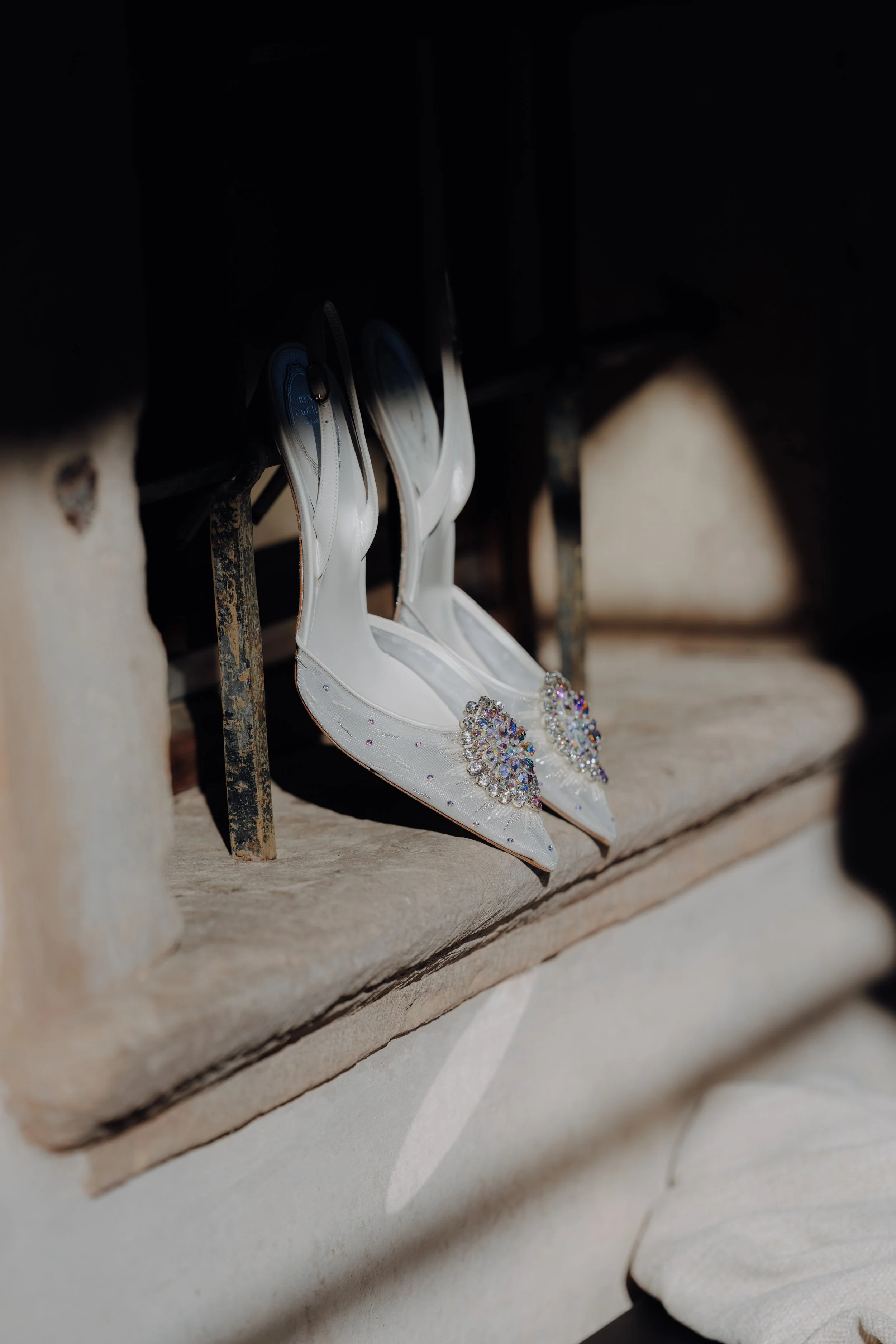 Weiße Brautschuhe mit Verzierungen, hängen an einer Metallstange in einem dunklen Raum.