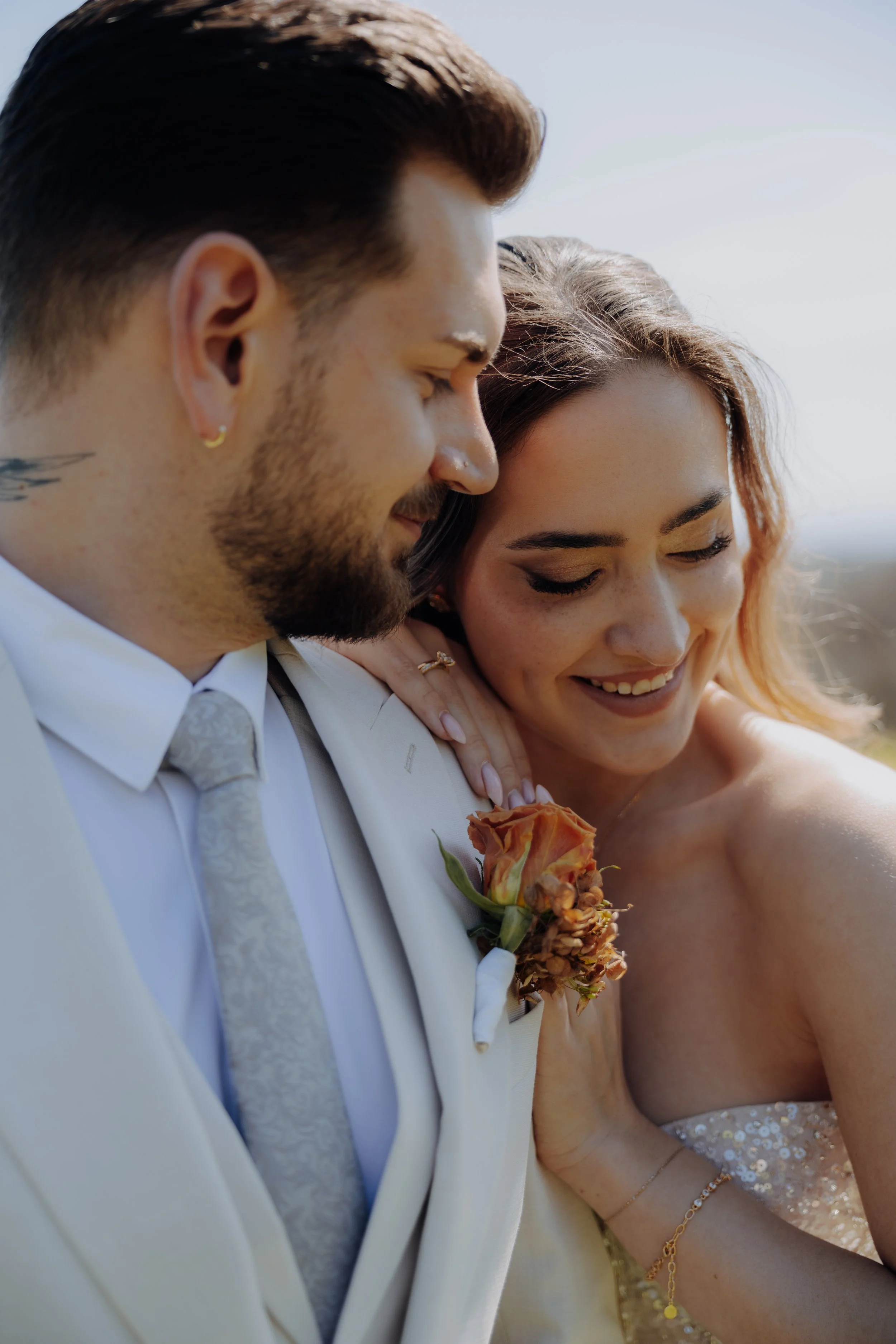 Ein Mann und eine Frau bei einer Hochzeitsfeier, die eng umarmt und lächelnd sind, mit dem Mann in einem weißen Anzug und der Frau in einem silbernen, sequenzierten Kleid, die Frau trägt Schmuck und hält einen Blumenstrauß.