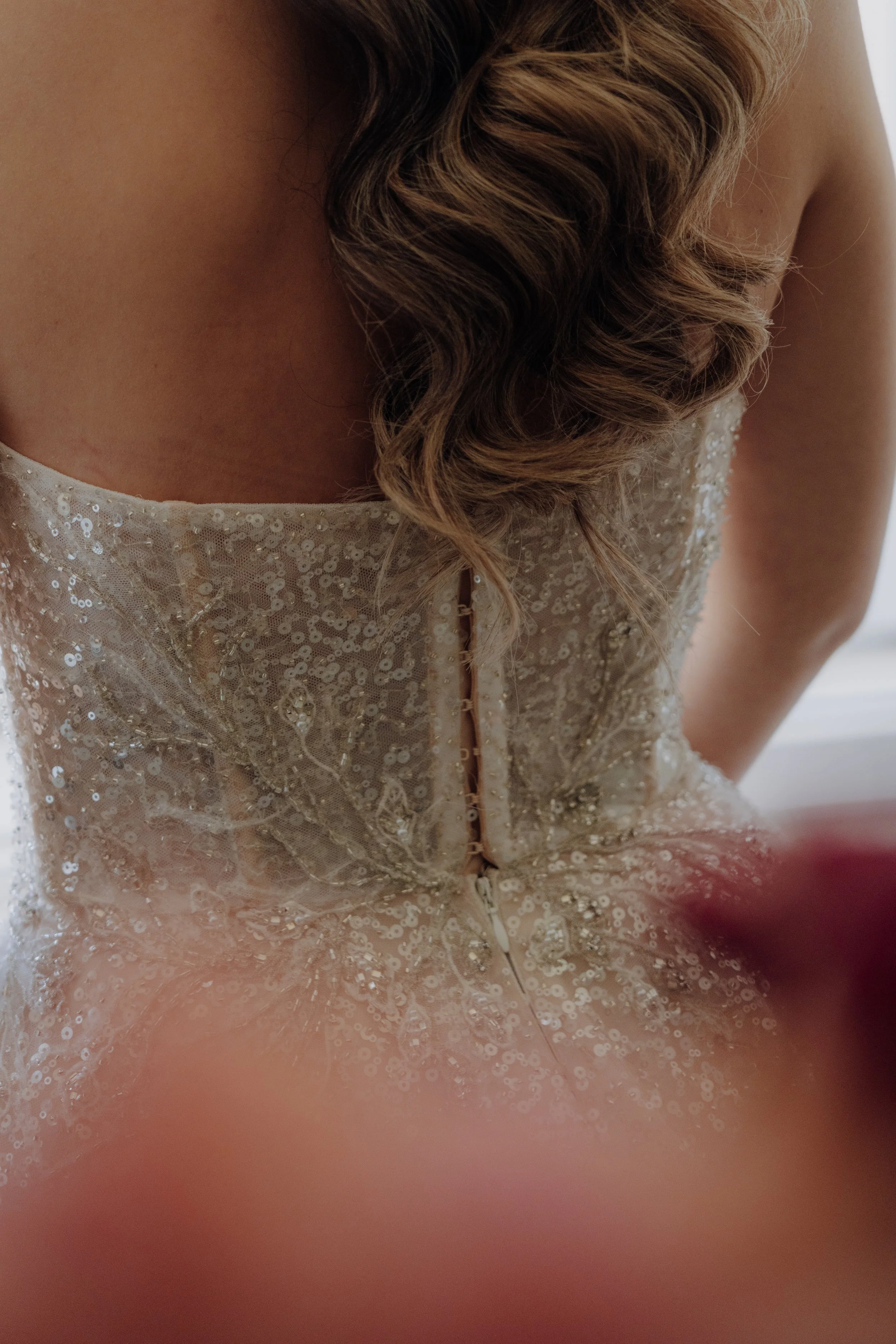 Nahaufnahme eines Brautkleides mit Pailletten und einer Zierde am Rücken. Haarstyling mit lockigem, braunem Haar. 