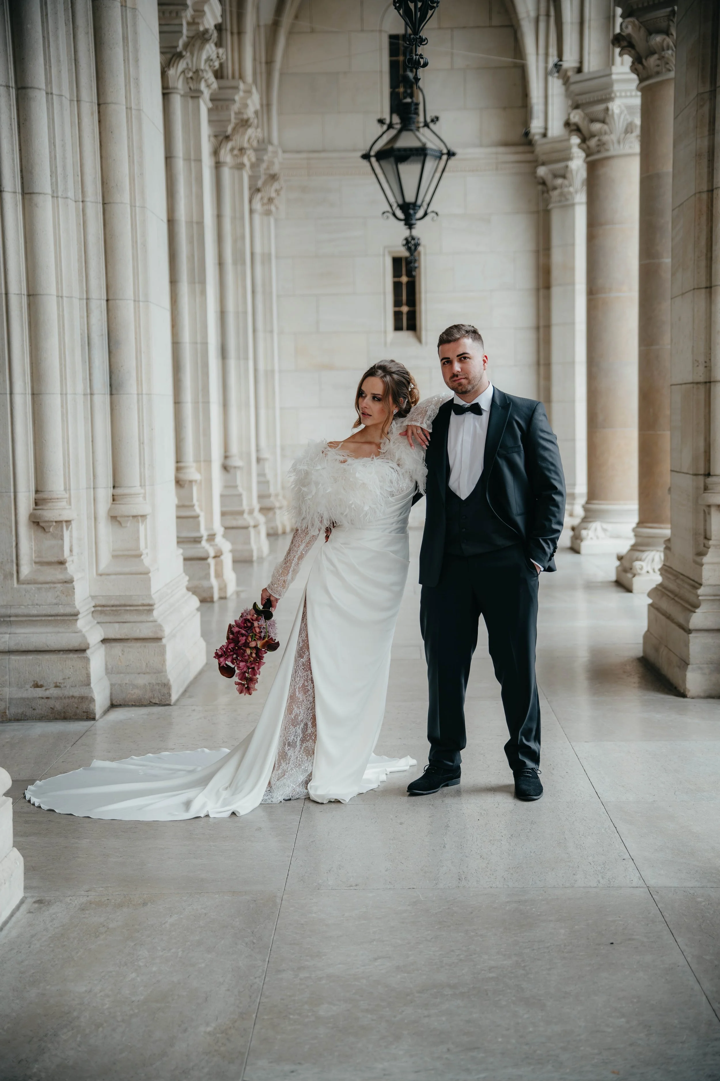 Ein Brautpaar in formeller Kleidung steht in einer eleganten, historischen Halle mit hohen Säulen und einer großen Pendelleuchte. Die Braut trägt ein langes, elegantes weißes Hochzeitskleid mit Perlen und Federn, hält einen Bouquet in der Hand und ha