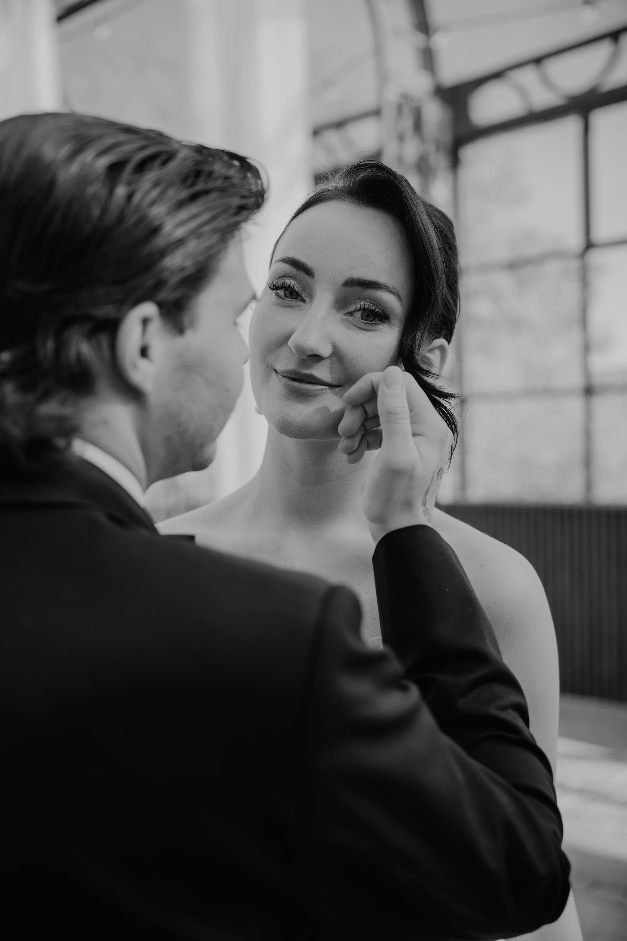 Ein Mann richtet Make-up bei einer Frau her, die lächelnd in die Kamera schaut, im Hintergrund ein großes Fenster.