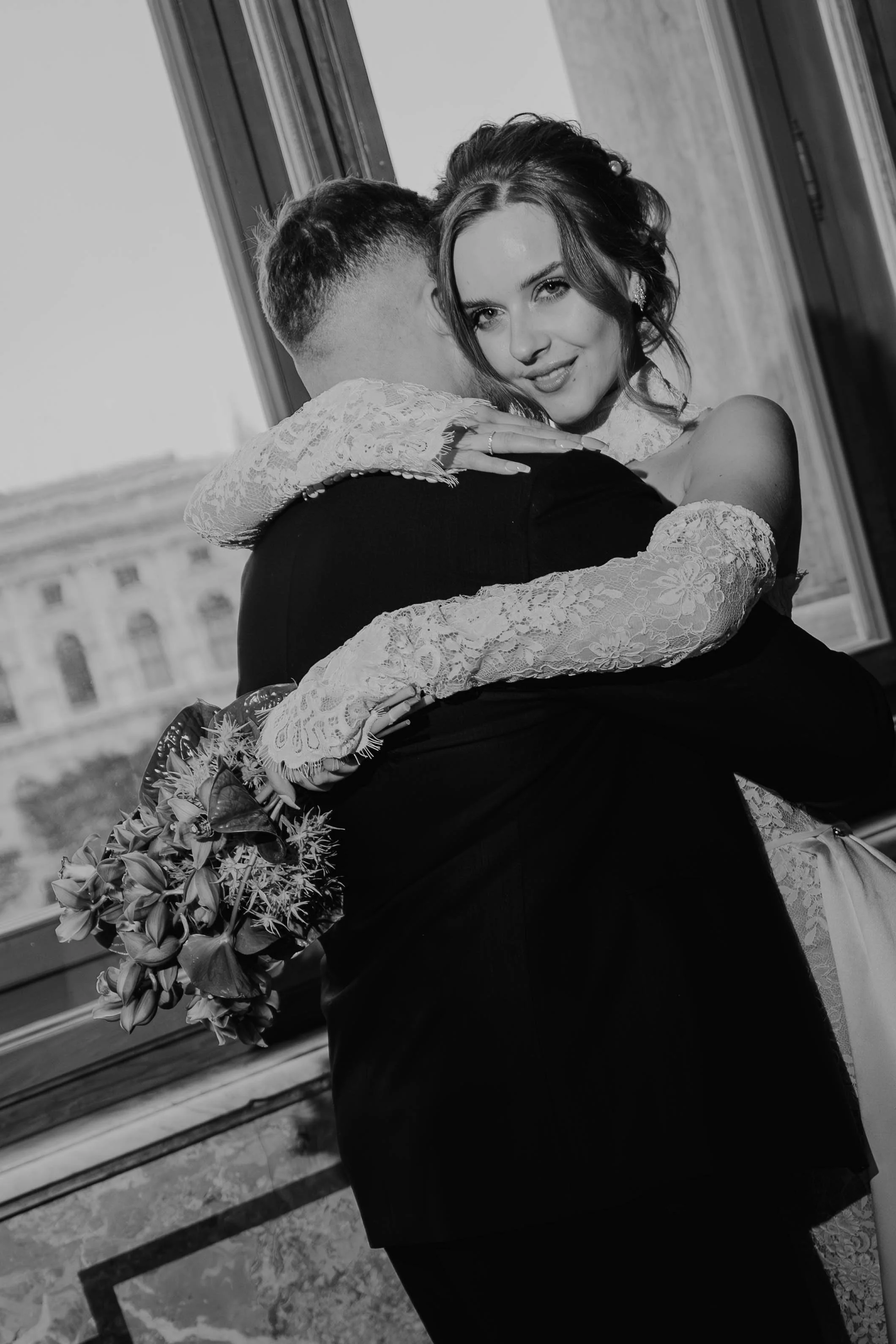 Ein Brautpaar beim Umarmen, das brautkleid und der Anzug sichtbar, das Paar steht vor einem Fenster mit Stadtblick, Schwarz-Weiß-Foto.