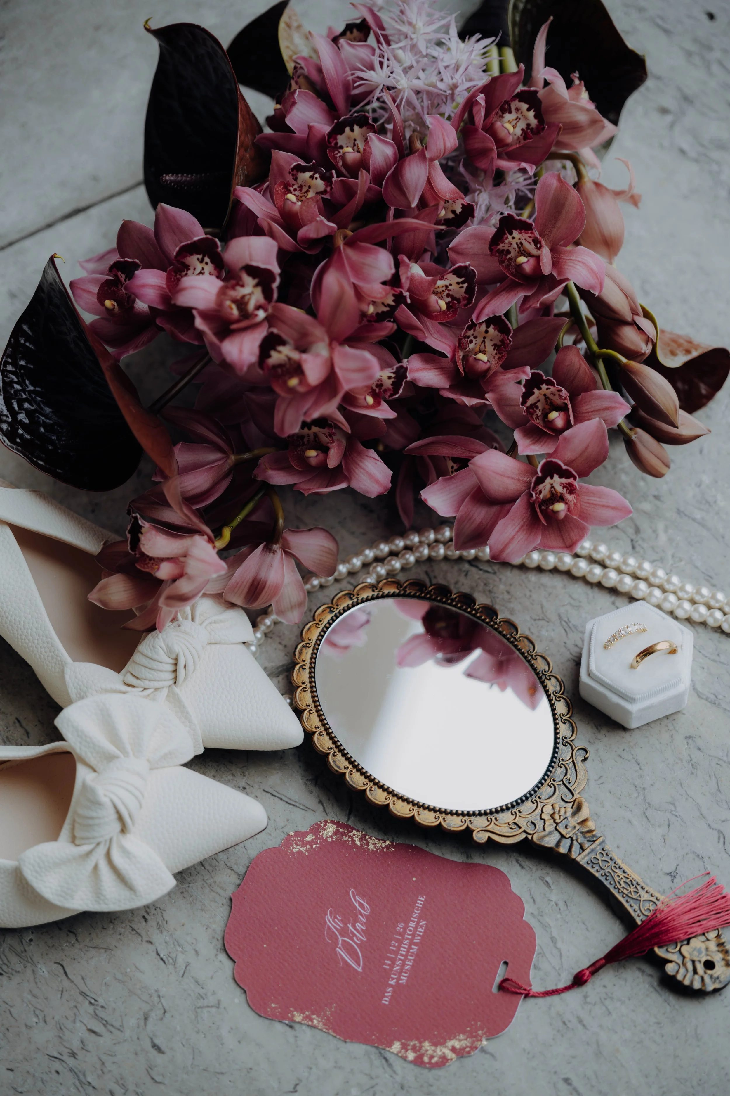 Ein Brautstrauß mit pinken und dunklen Blumen, eine silberne handliche Handspiegel, eine weiße Schleife mit Schleifenknoten, eine rosa Einladungskarte mit Goldrand und goldenem Text, sowie Schmuckstücke, auf einer Steinoberfläche.