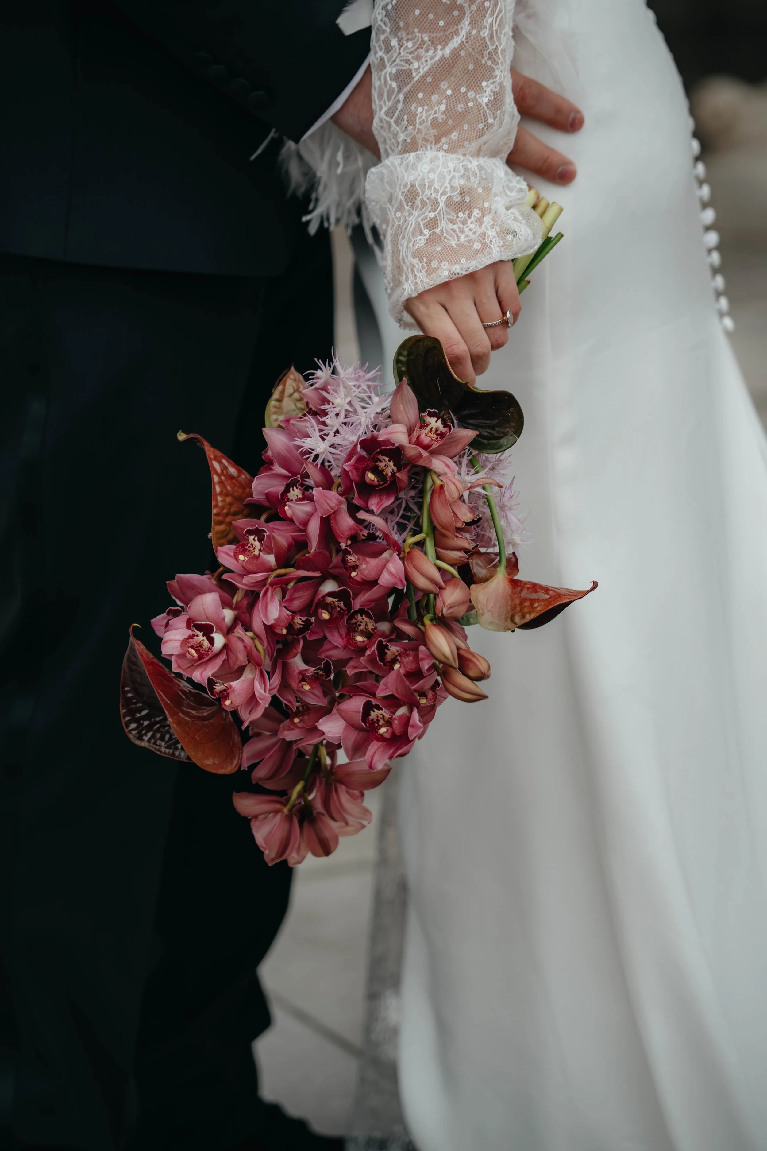 Ein Brautpaar hält sich bei einer Hochzeit an den Händen, die Braut trägt eine weiße Hochzeitssilhouette und einen Ring, die Braut trägt ein langärmeliges Brautkleid mit Spitze. Die Braut hält einen bunten Blumenstrauß aus pinken, weißen und roten Bl