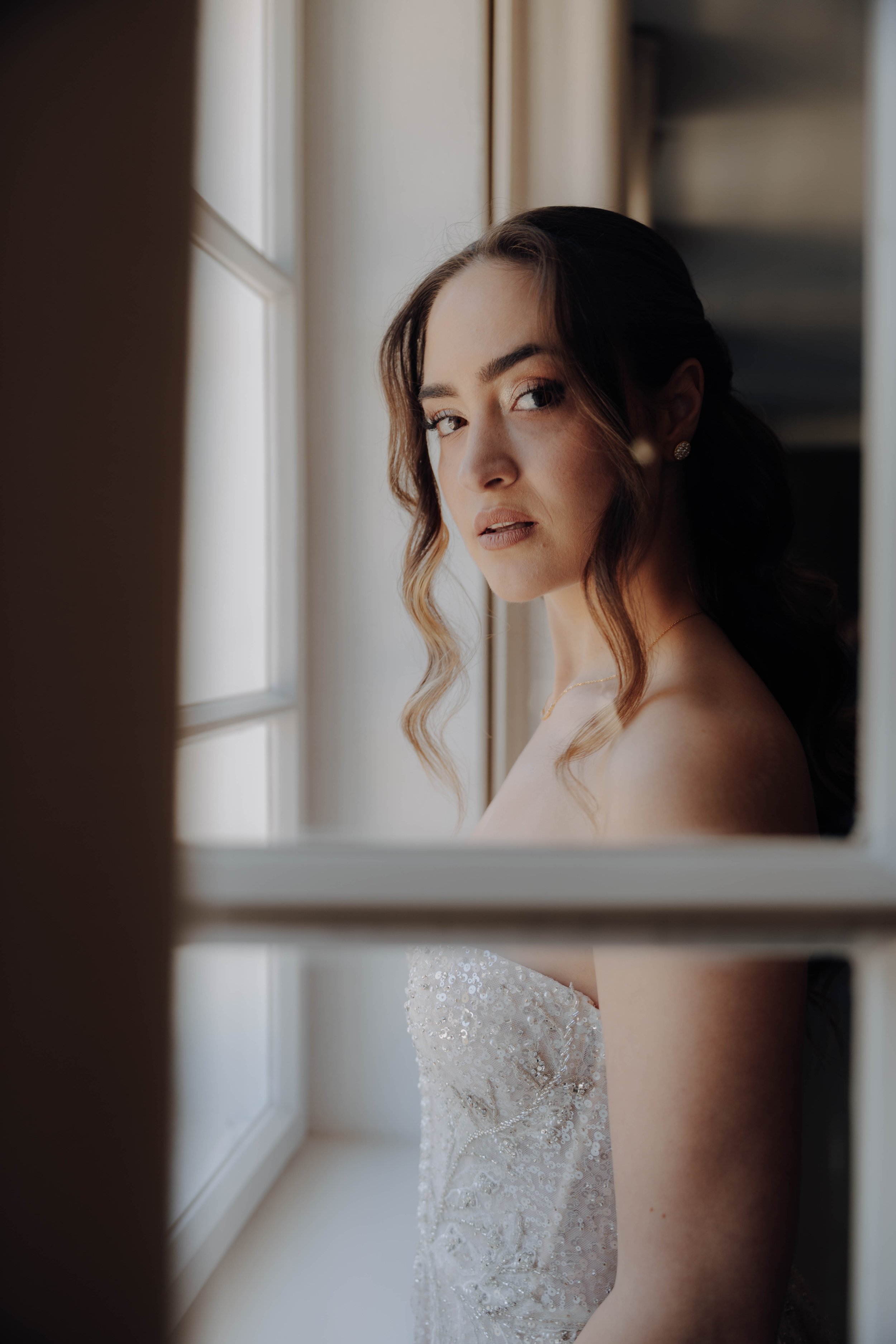 Eine Frau, vermutlich eine Braut, blickt durch ein Fenster. Sie trägt ein weißes, elegante Brautkleid mit Pailletten. Ihre Haare sind lockig und sie trägt Ohrringe.