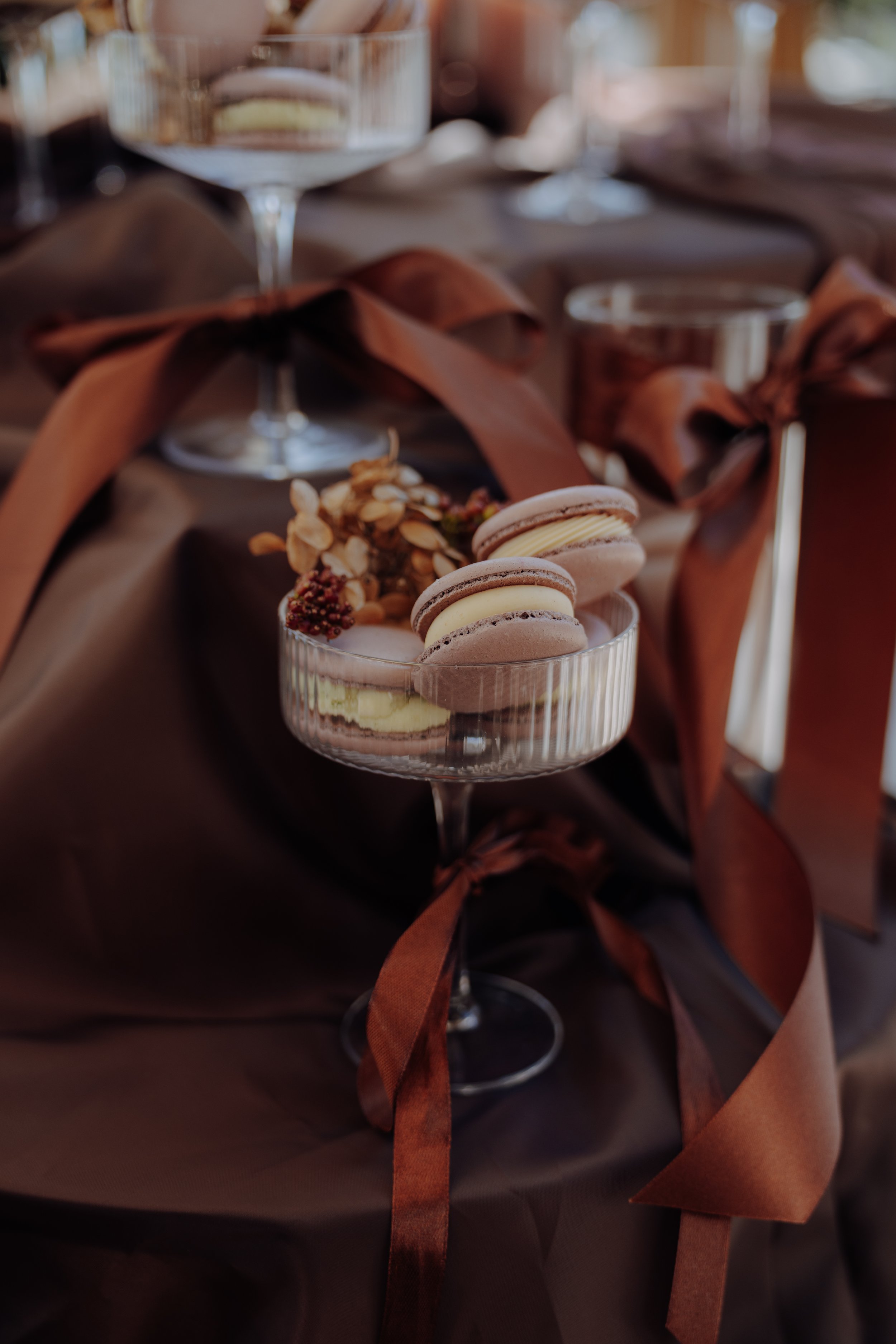 Ein Glas mit mehreren Macarons und einer kleinen Blumen- und Nussdekoration, umgeben von braunen Bändern, auf einem festlich gedeckten Tisch im Hintergrund