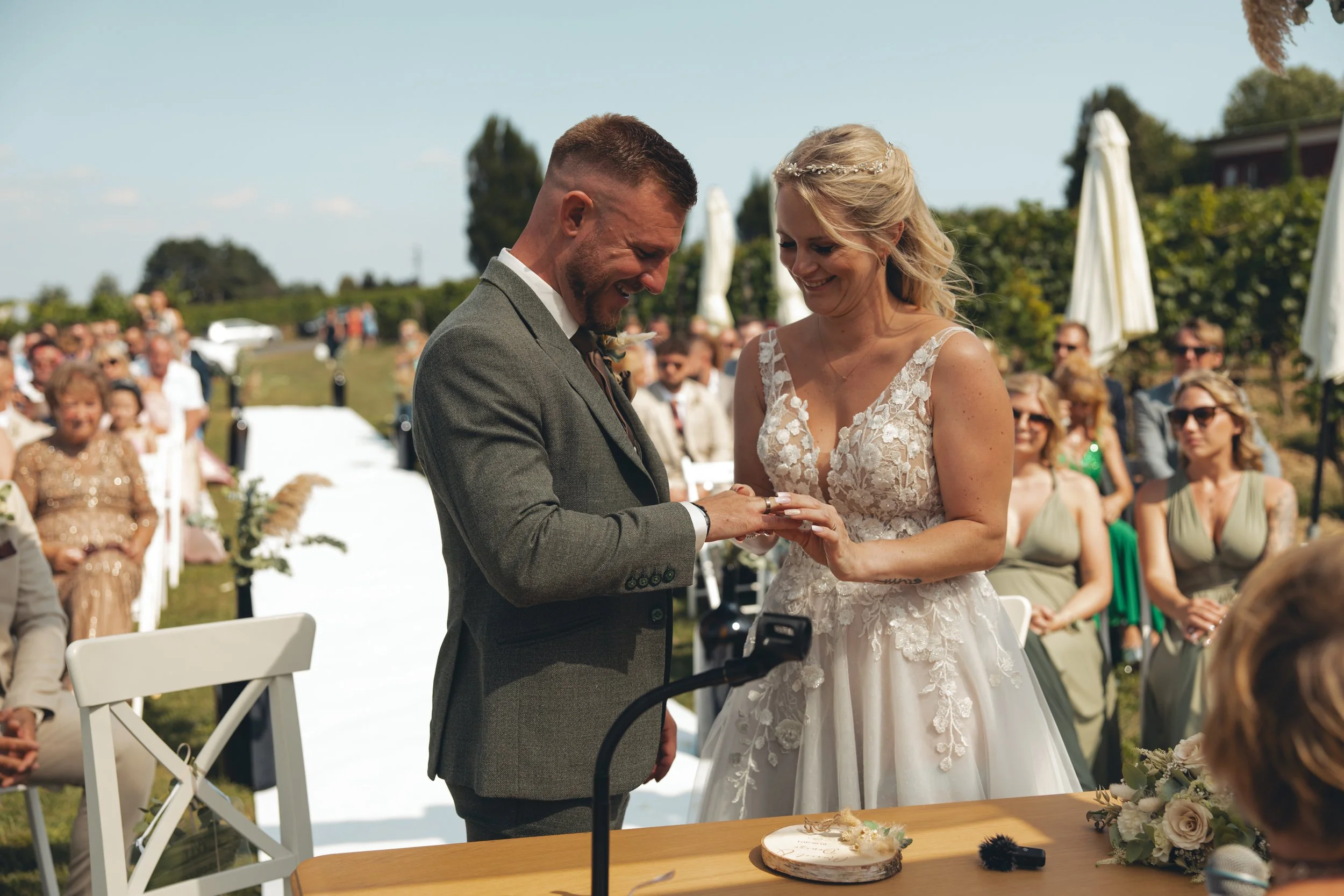 Ein dafür lächelndes Brautpaar während ihrer Hochzeitszeremonie im Freien, umgeben von Gästen im Sonnenschein.