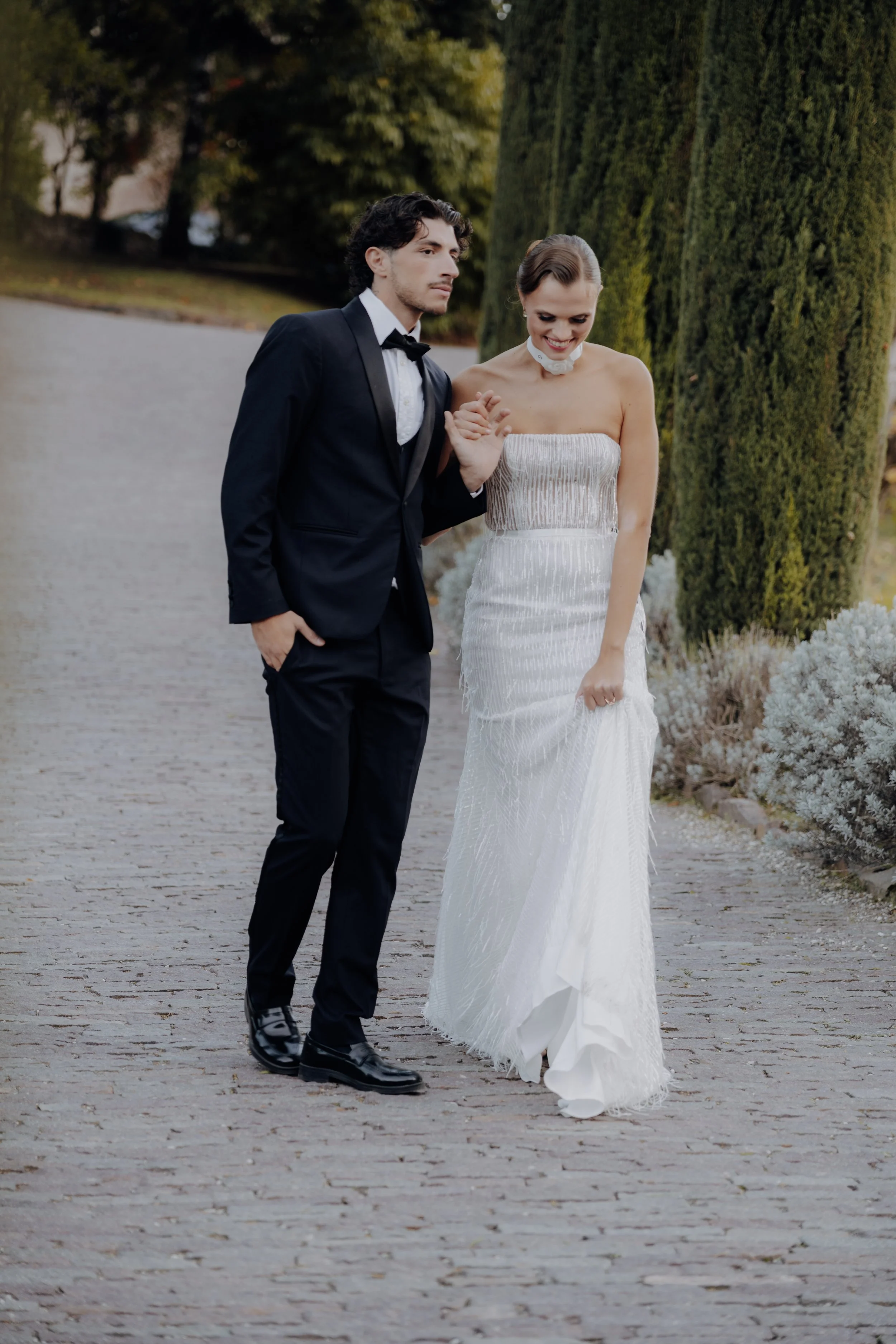 Ein Mann in einem schwarzen Anzug mit Fliege hält eine Frau in einem weißen Brautkleid an der Hand, während sie auf einem Gehweg im Freien gehen.