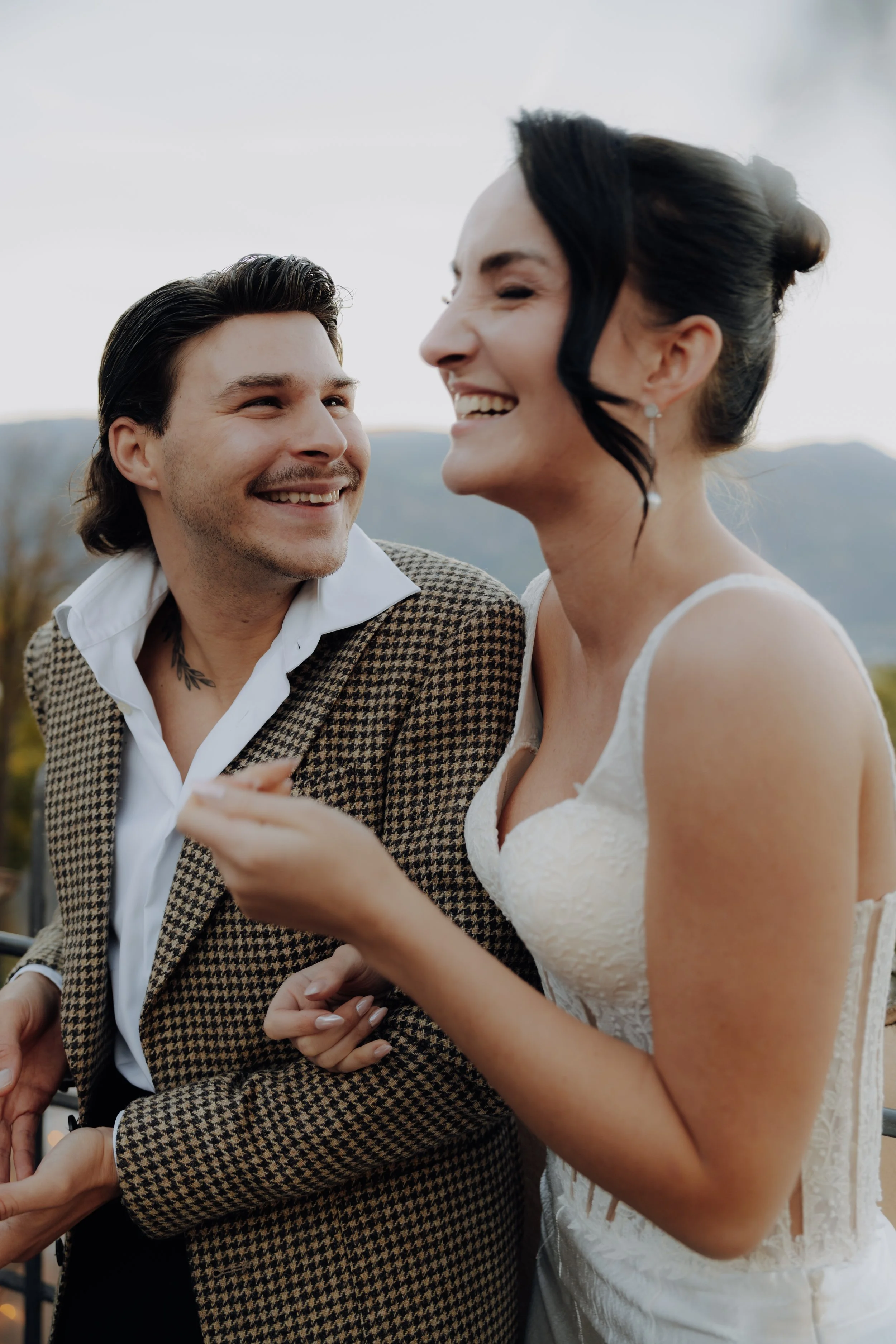 Ein Mann und eine Frau beim Lachen, nahe beieinander, bei einem Outdoor-Event oder einer Feier. Der Mann trägt einen karierten Anzug und ein weißes Hemd, die Frau trägt ein weißes Spitzenkleid, möglicherweise eine Braut. Es herrscht eine fröhliche At