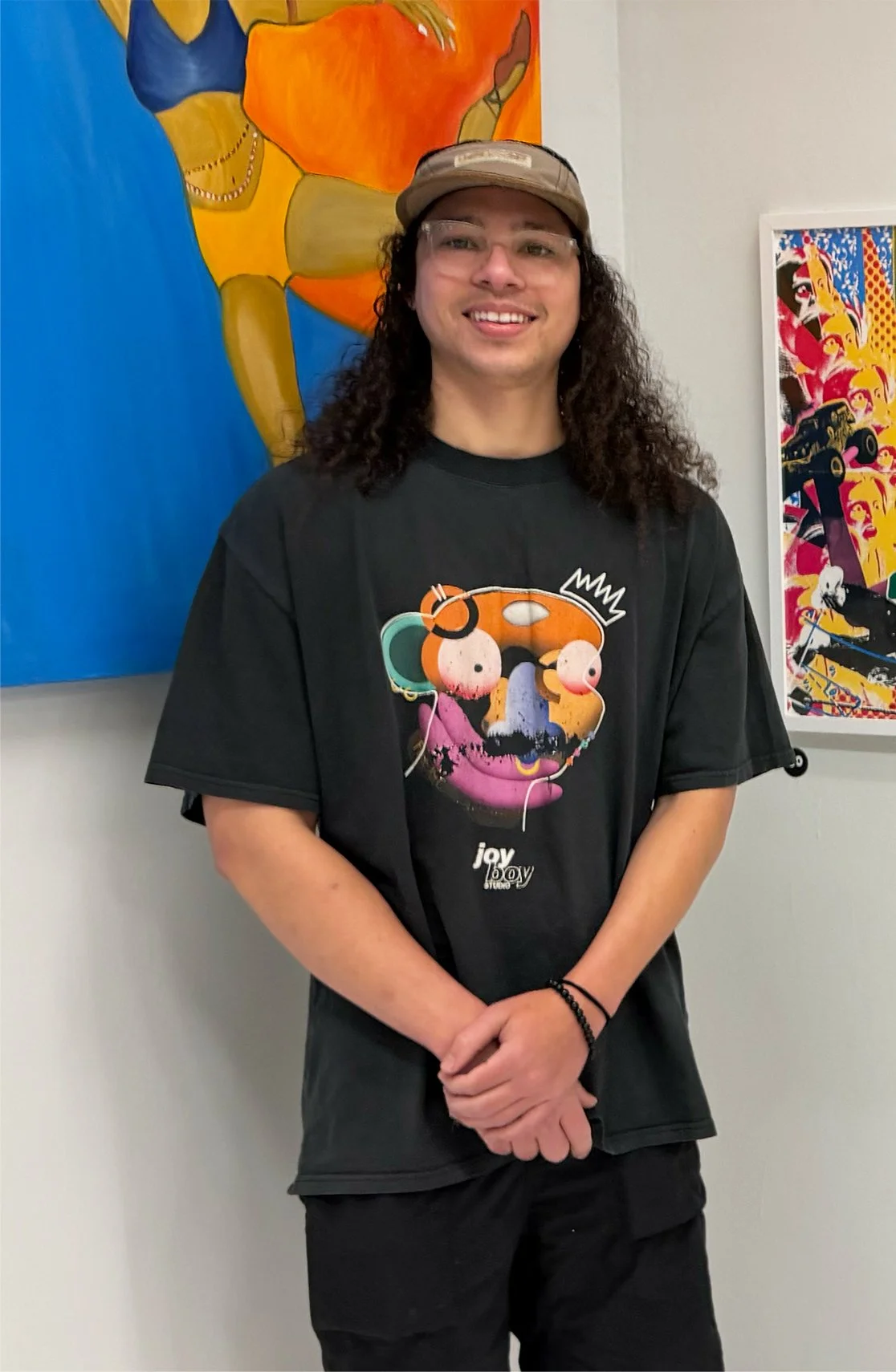 A person with long curly dark hair, glasses, and a brown cap, smiling, standing in front of colorful abstract paintings. They are wearing a black T-shirt with a graphic of a stylized face and the words 'joy 15 joy' on it.
