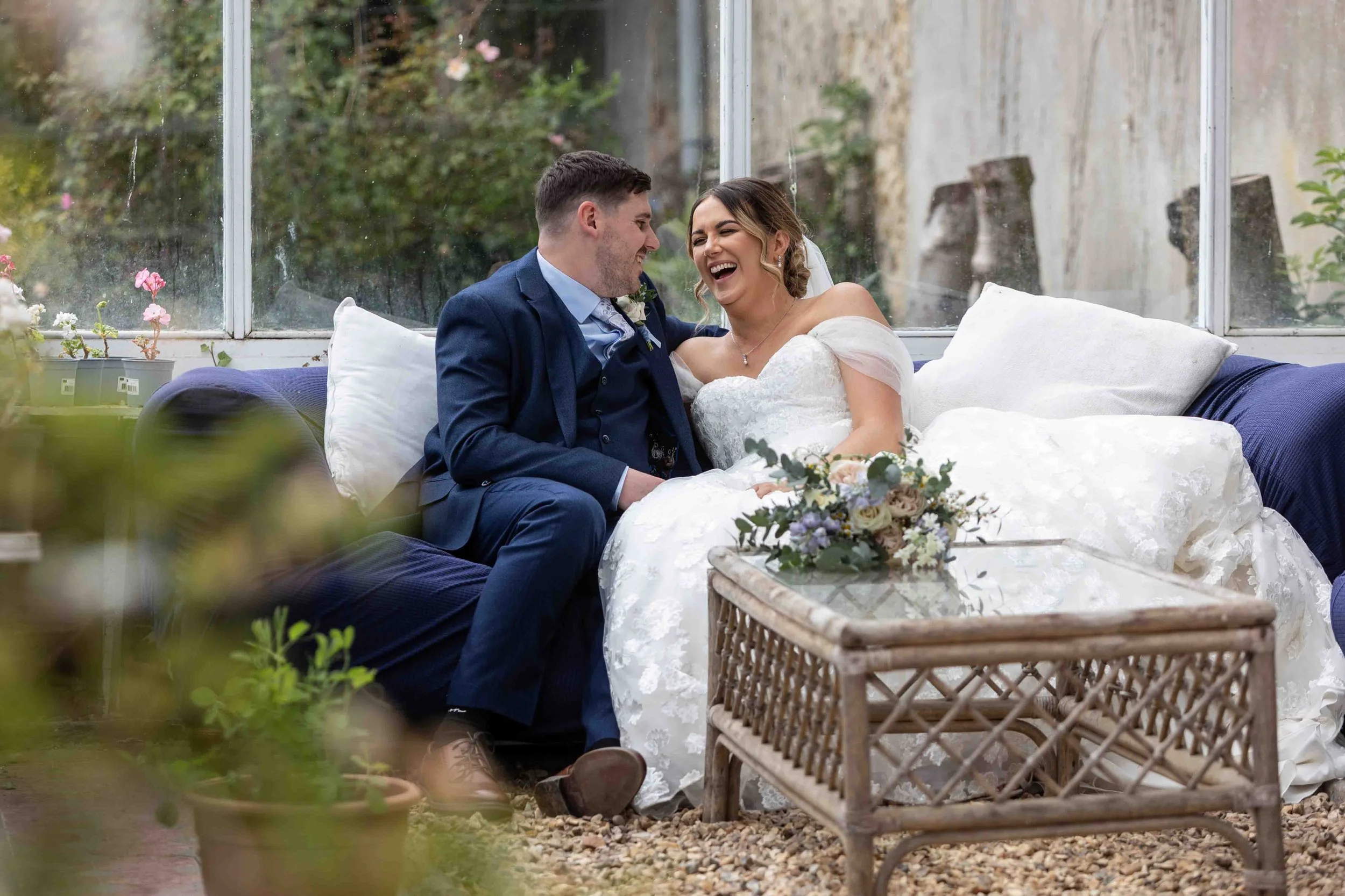 A newlywed couple sitting on a couch inside a glass-enclosed garden or conservatory, laughing and smiling at each other. The bride is wearing a white wedding dress with lace details, and the groom is dressed in a dark blue suit. There are potted plants and flowers around them, with a floral bouquet on a wicker table in front. The background shows plants and a stone wall outside the glass enclosure.