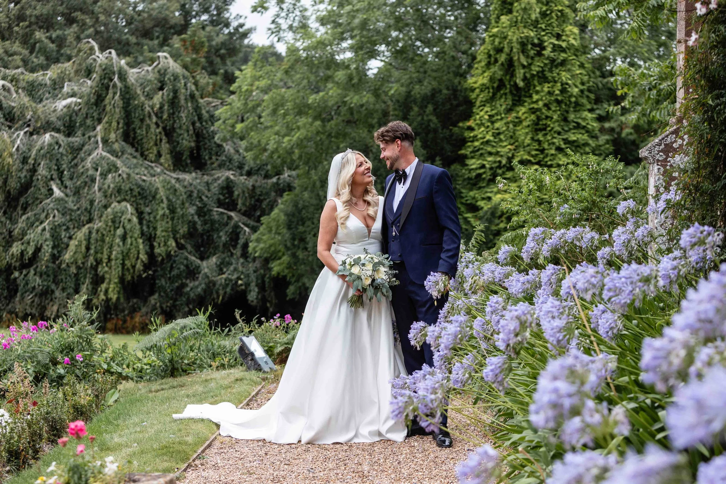 A bride and groom standing together in a garden, surrounded by greenery and purple flowers, smiling at each other.