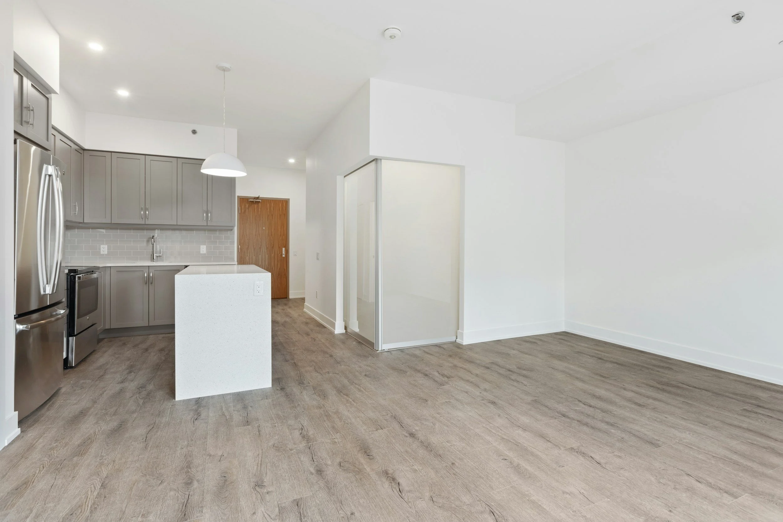 Empty modern kitchen with gray cabinets, white countertops, stainless steel appliances, a wooden door, glass sliding door, and wood flooring.