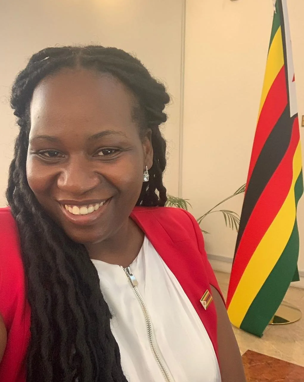 A woman with long dreadlocks smiling, wearing a red blazer over a white top, standing in front of a Zimbabwe flag.