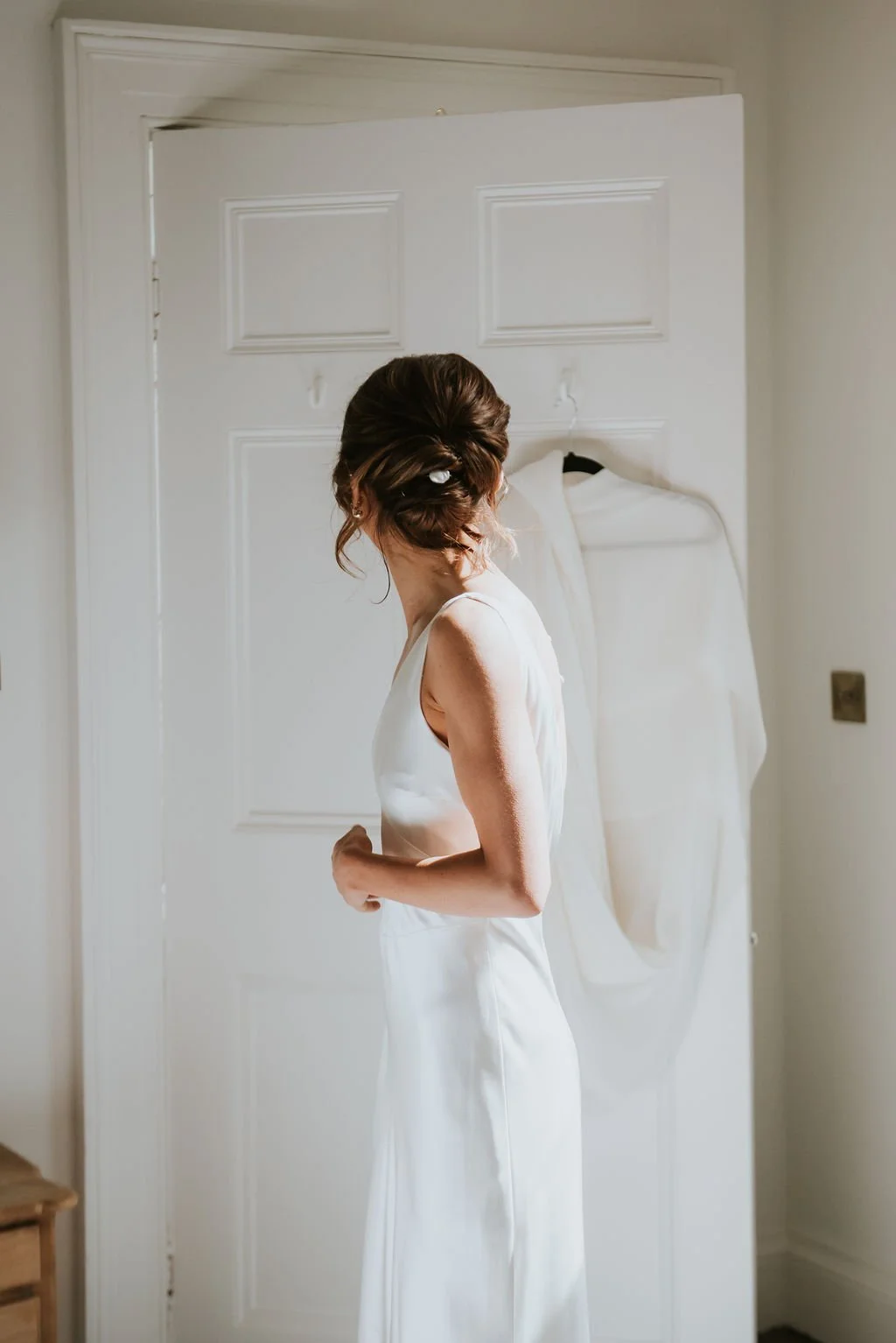 A woman in a white satin dress standing in front of a white door, with her back to the camera, her hair styled in an updo.