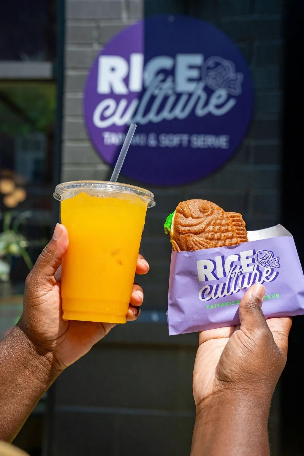 A person holding a cup of yellow mango juice and another person holding a fish-shaped pastry in a purple paper wrapper with 'RICE & Culture' logo, in front of a sign that says 'RICE & Culture, Taiyaki & Soft Serve.'