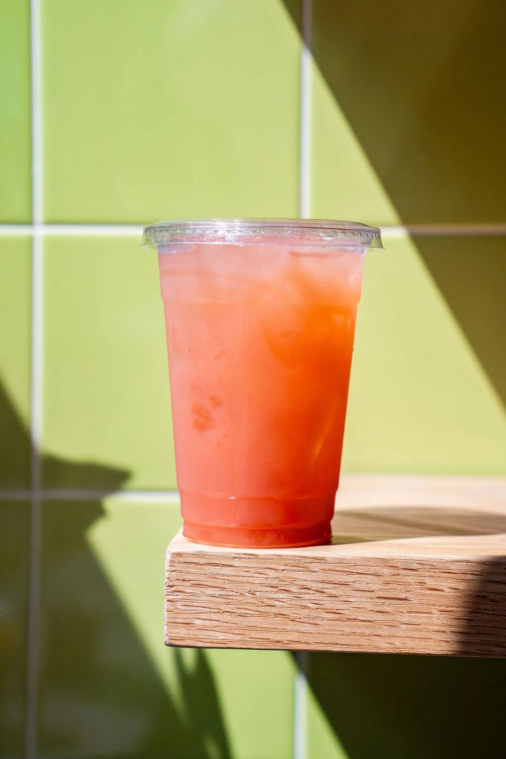 A clear plastic cup filled with pink iced beverage, placed on a wooden surface against a green tiled wall with sunlight casting shadows.