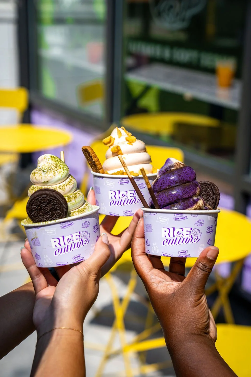 Three people holding cups of ice cream with toppings on a sunny day at an outdoor seating area with yellow chairs in the background.