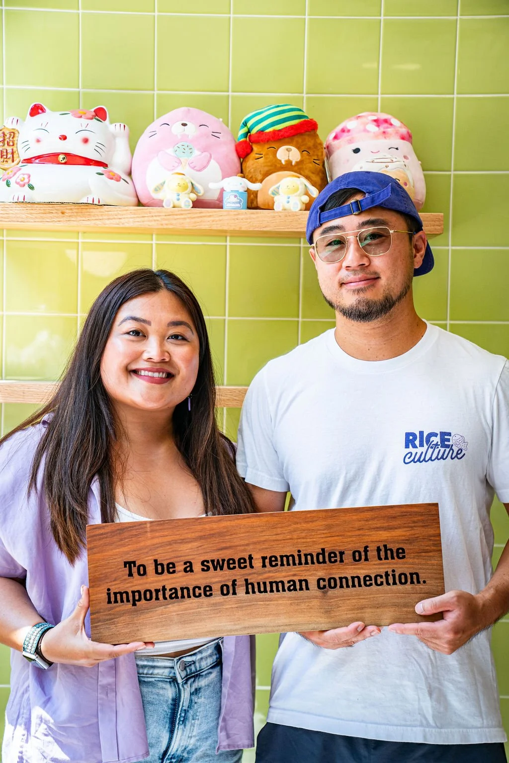 A woman and a man smiling and holding a wooden sign that says 'To be a sweet reminder of the importance of human connection.' They are standing in front of a green tiled wall with colorful plush toys on a shelf above their heads.