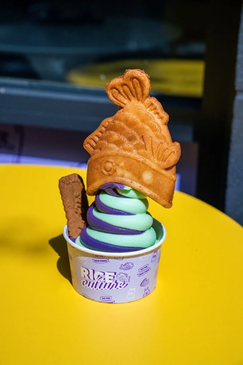 Colorful swirl soft serve ice cream in a cup, topped with a fish-shaped cookie and a fish-shaped waffle biscuit, on a yellow surface.