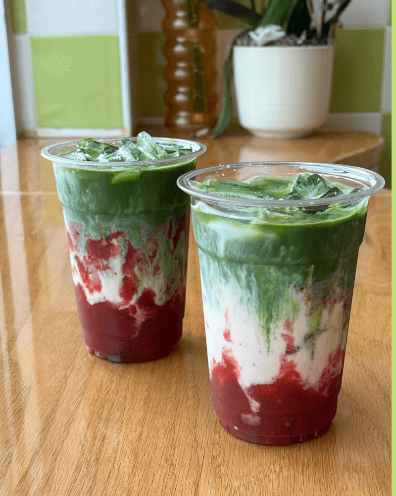 Two plastic cups filled with a layered drink containing red syrup at the bottom, white creamy layer, and green topping, with ice on top, placed on a wooden table.