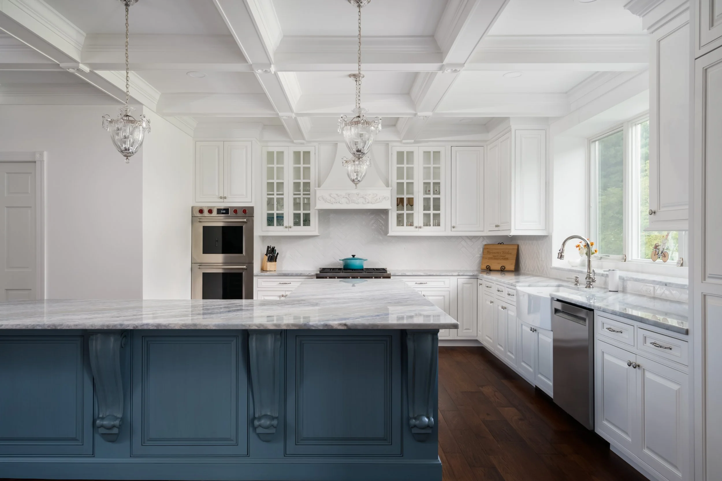 A luxury white kitchen with glass-front cabinets, a large marble island with blue detailing, and coffered ceilings with crystal pendant lights.