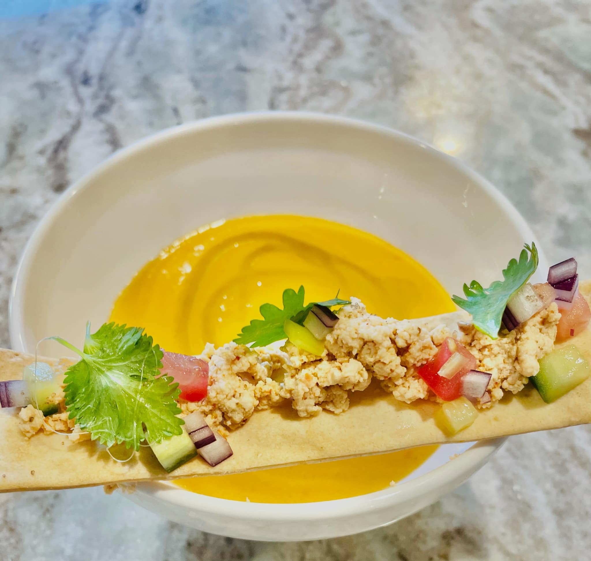 A bowl of creamy yellow soup garnished with chopped cucumbers, tomatoes, purple onions, cilantro, and a crunchy breadstick or cracker on the side.