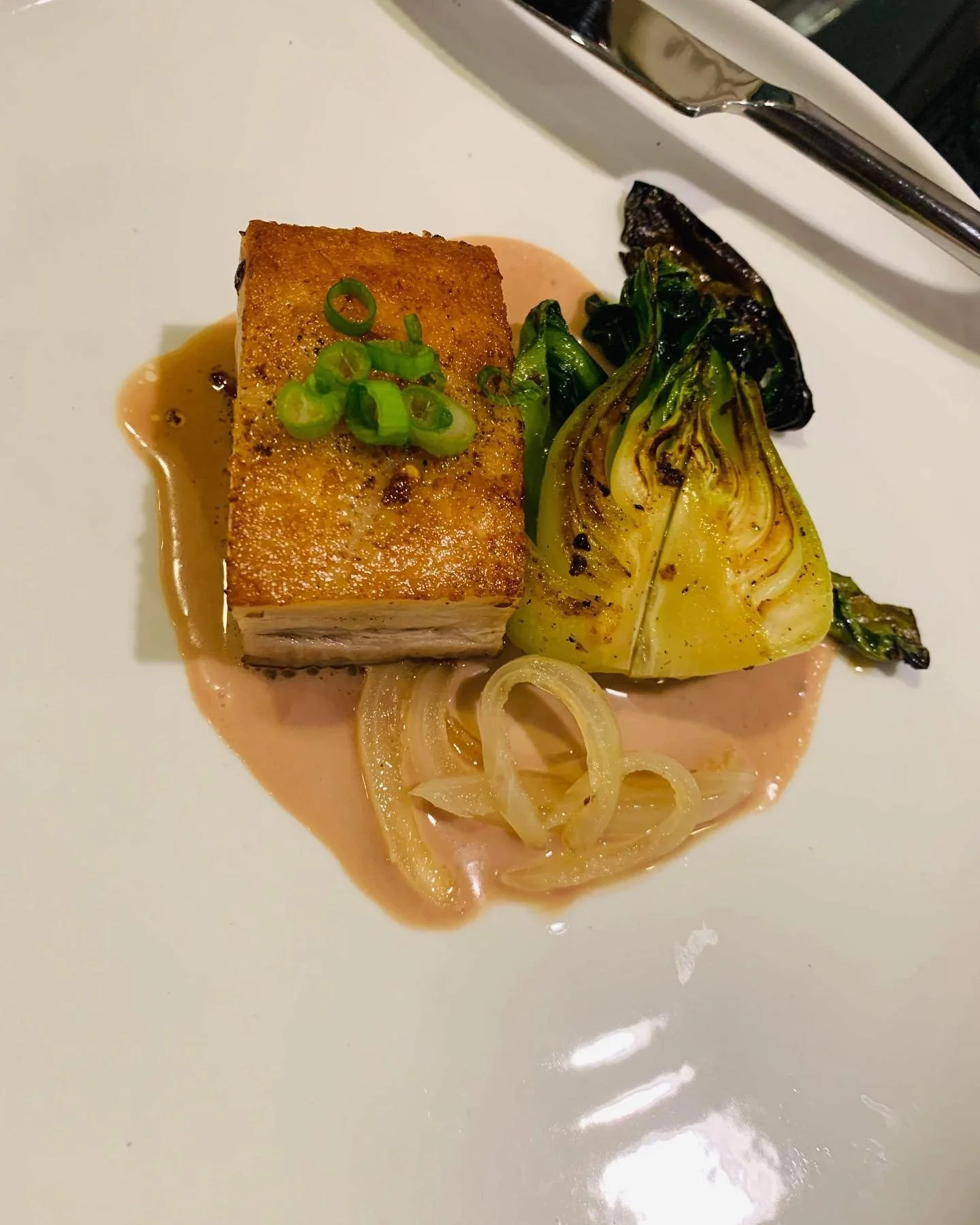 A plated dish featuring a crispy piece of fish topped with chopped green onions, served with grilled fennel and vegetables on a pink sauce.