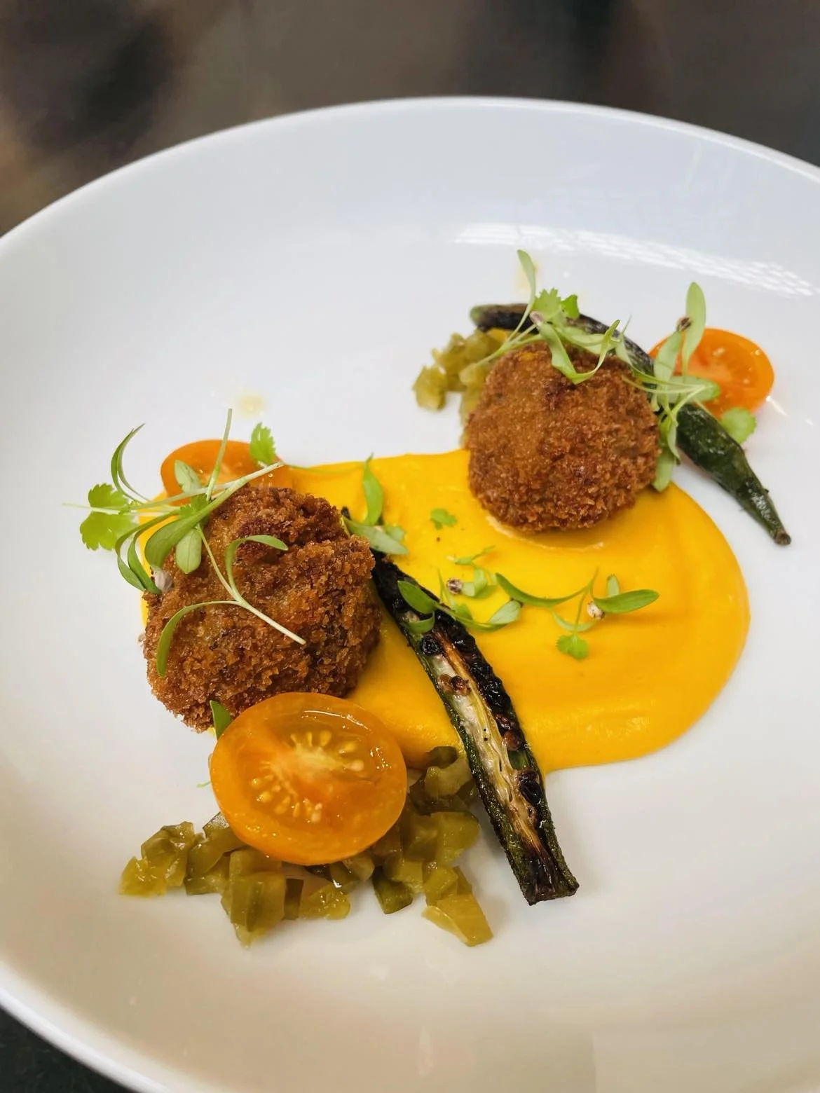 Plate with two breaded and fried croquettes, yellow sauce, grilled green chili peppers, cherry tomatoes, and microgreens.
