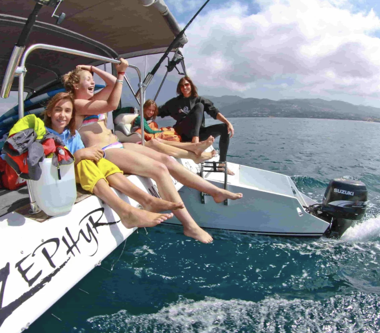 Four people smile and dangle their feet off Zephyr after surf lessons, enjoying the ocean under a partly cloudy sky.