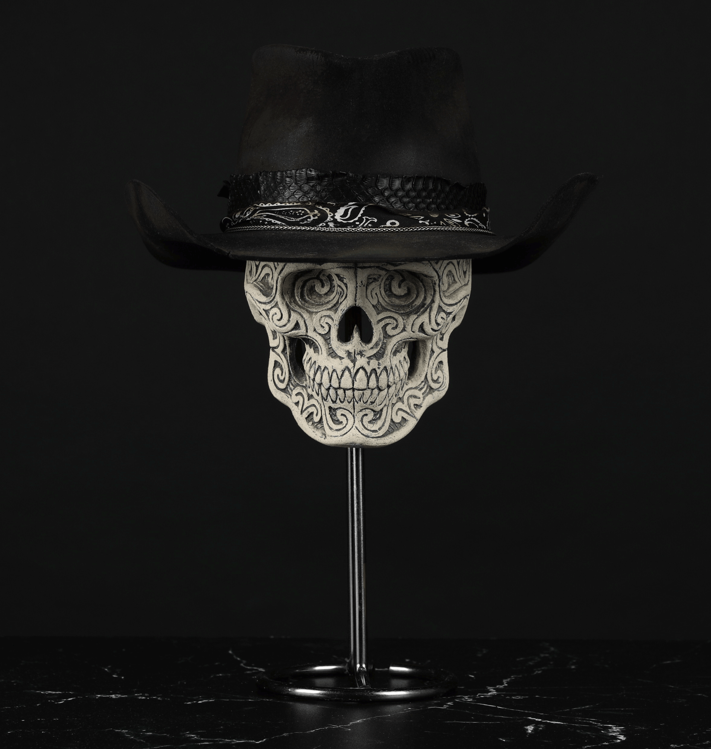 A black cowboy hat with a band, placed on top of a decorated skull with a spike in the base, on a dark surface with a black background.