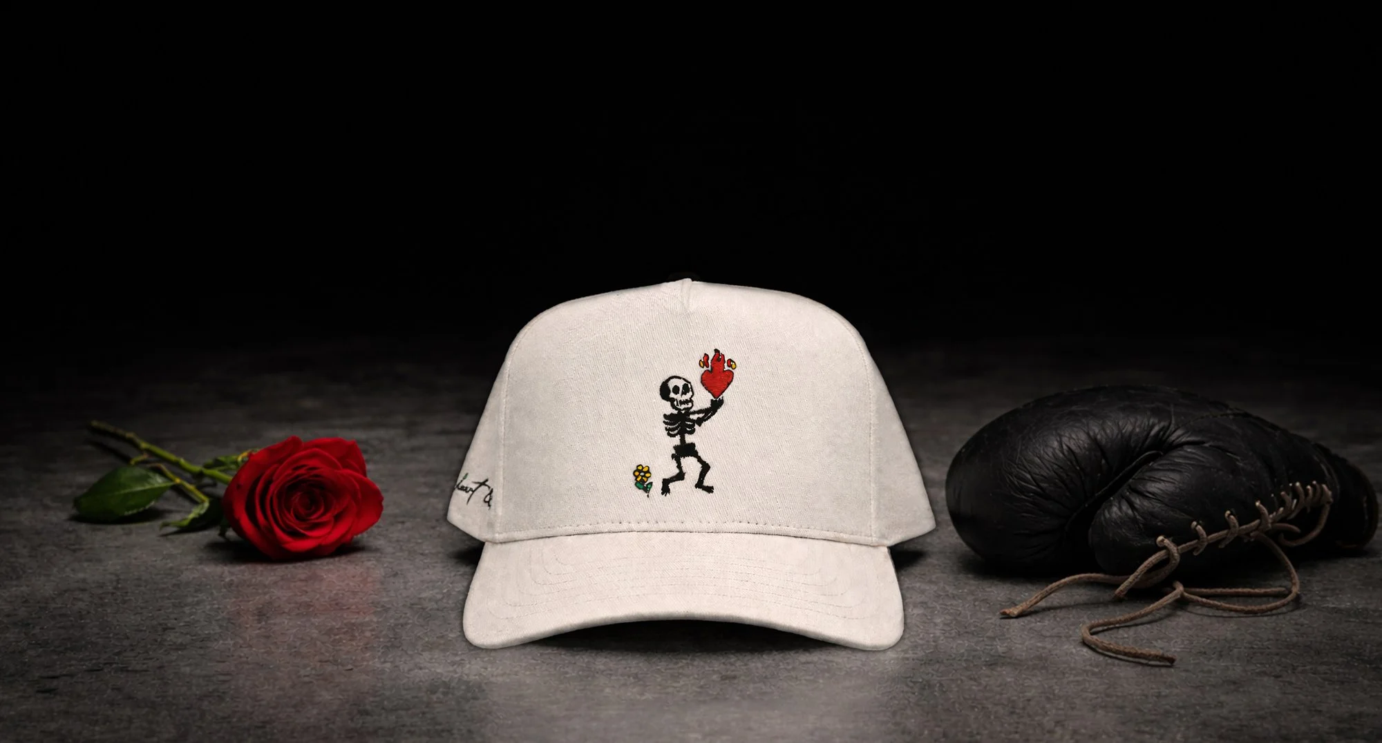 White baseball cap with a skeleton illustration holding a burning heart, a red rose on the left, and a black baseball glove on the right, against a dark background.