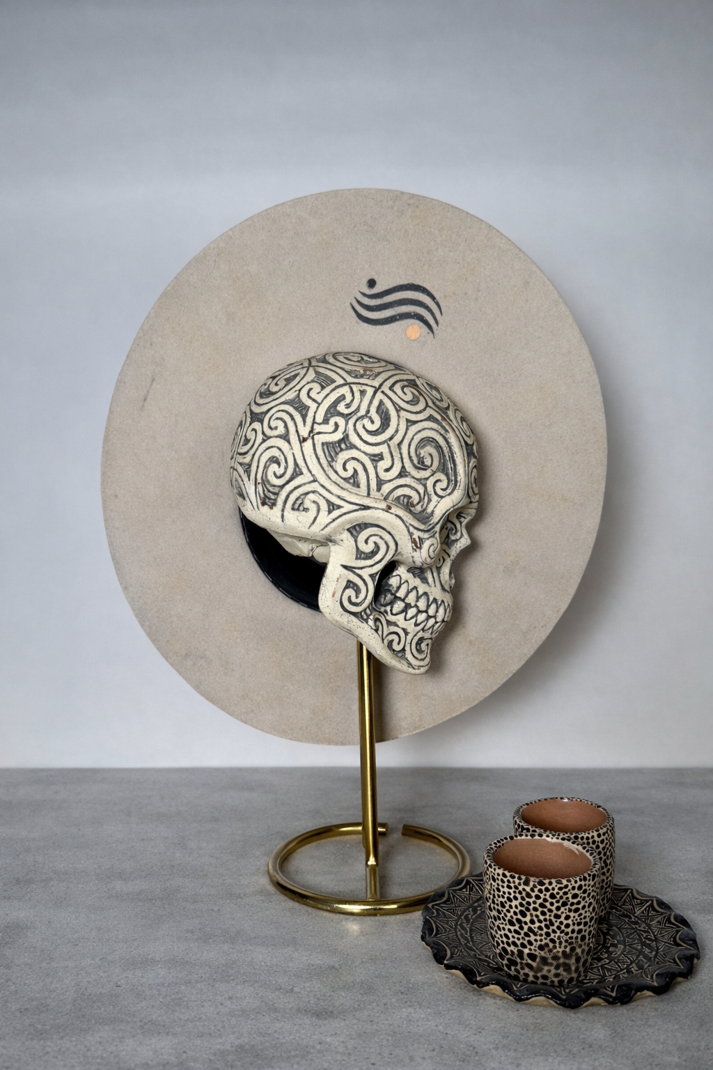 Decorative skull with black and white swirl patterns on a stand, with a textured backdrop, two cups with matching patterns on a tray, and a symbol of waves and a dot above.