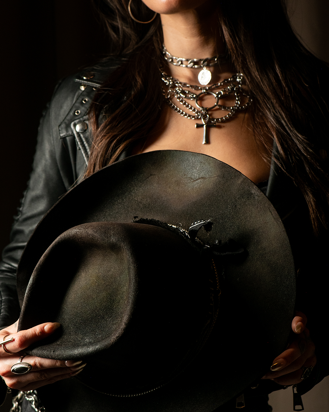 A woman holding a cowboy hat, wearing layered silver necklaces, a leather jacket, hoop earrings, and rings.