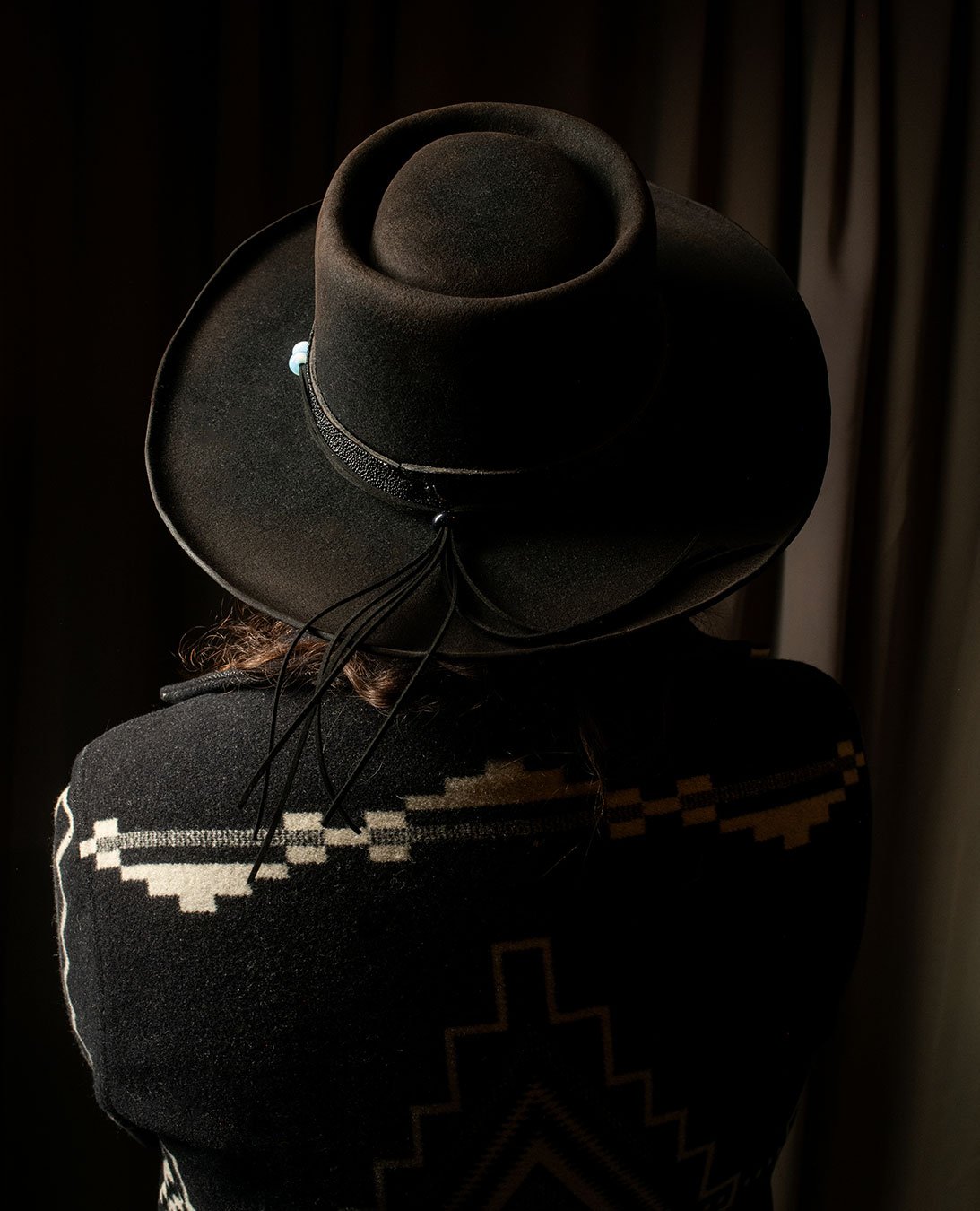 A person wearing a black hat with a wide brim and decorative string, seen from behind, in a dark setting.