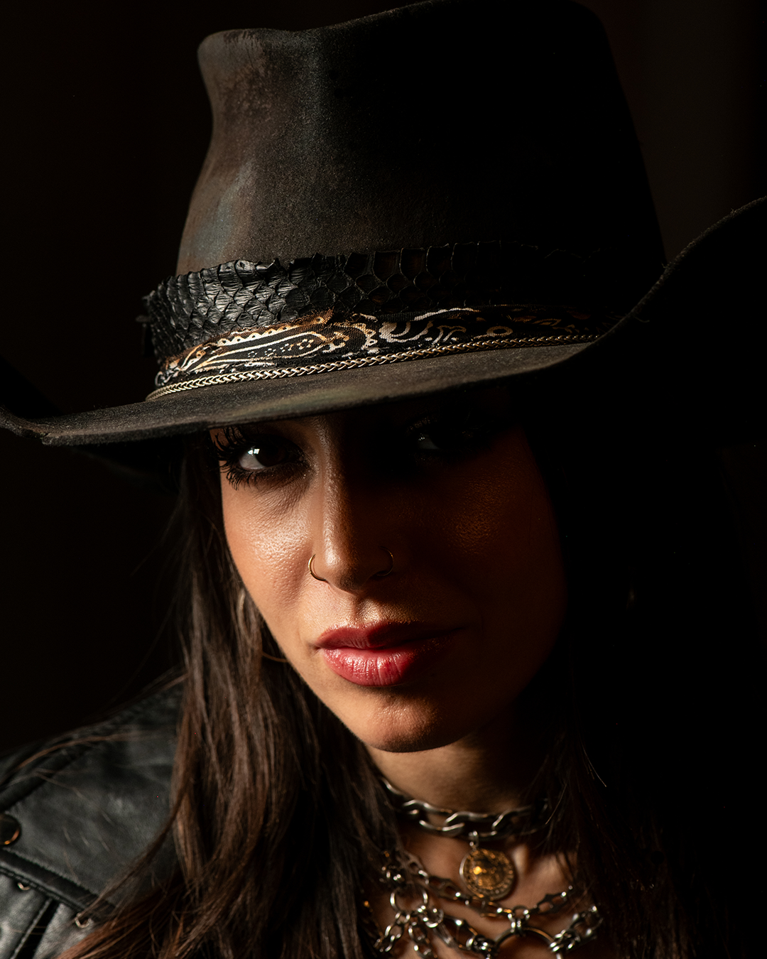 A woman wearing a dark cowboy hat with decorative bands, layered chains, and jewelry, with dark makeup, a lip piercing, and a leather jacket, looking directly at the camera in a dimly lit setting.