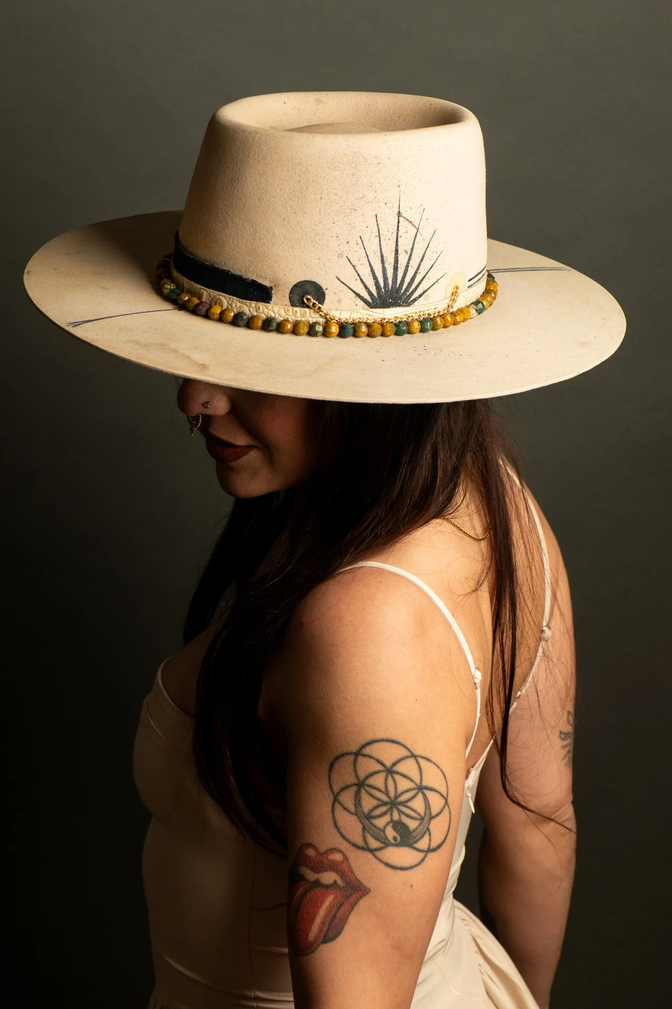 A woman wearing a large wide-brimmed hat with decorative beads and artwork, with long dark hair, tattoos on her arm, and a septum piercing, against a plain dark background.