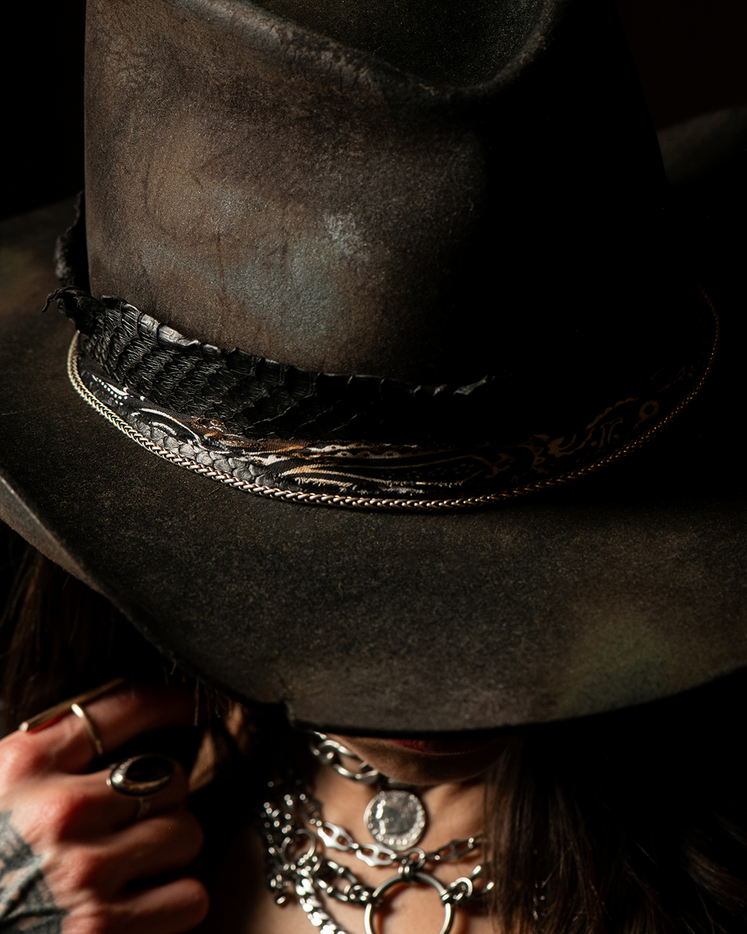 Close-up of a person wearing a black cowboy hat, layered silver jewelry, and a black fabric band around the hat. The person's face is partially obscured, with dark hair and rings visible on fingers.