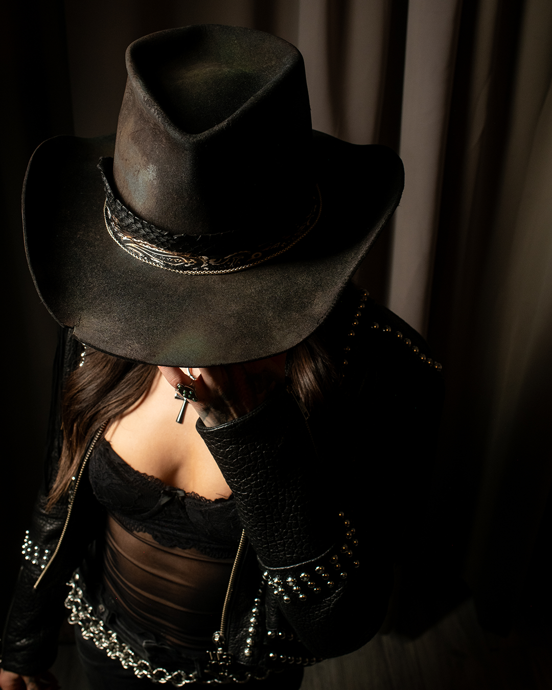 A person wearing a black cowboy hat, a black leather jacket with silver studs, a black lace top, and multiple silver chains, standing in front of a dark background.