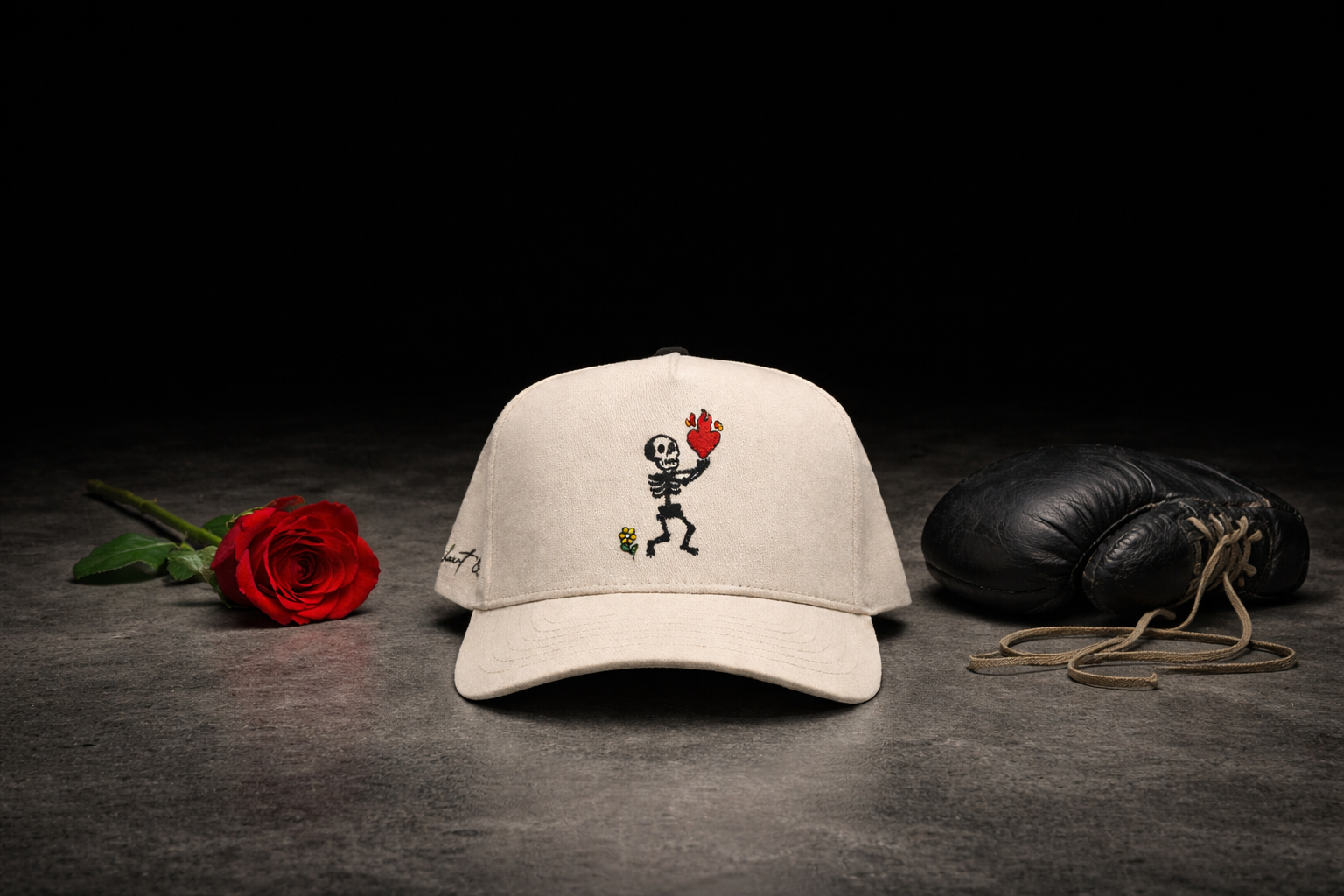 A beige cap with embroidered skeleton holding a flaming heart, a single red rose, and a vintage black boxing glove on a dark surface.