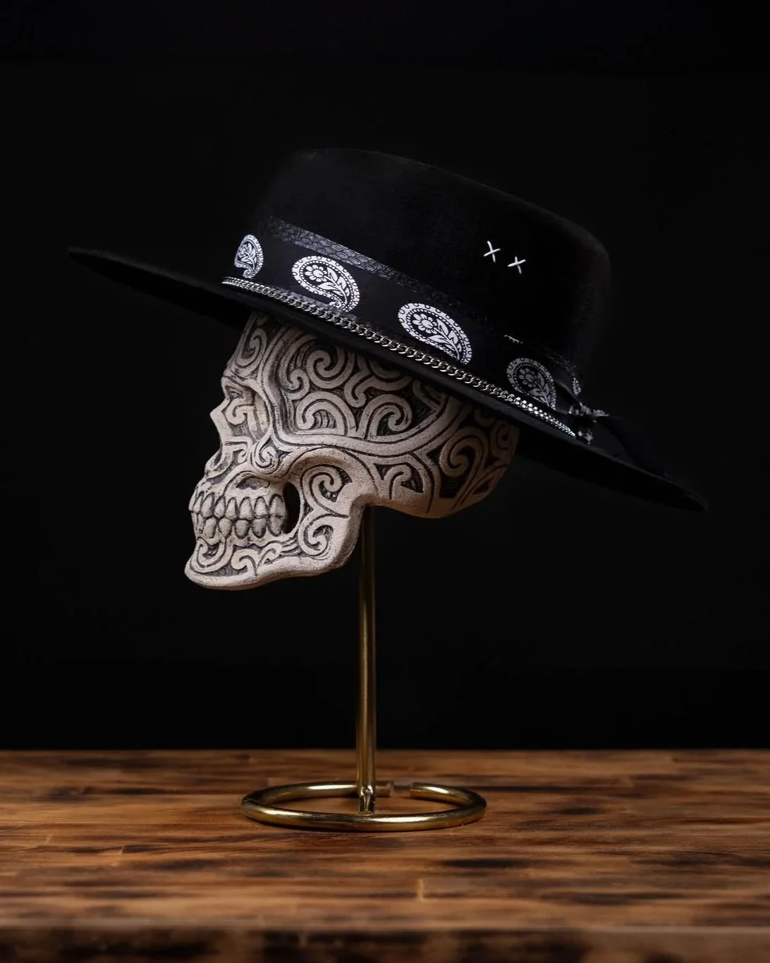Decorative skull sculpture wearing a black hat with paisley pattern and bandana against a black background, placed on a wooden surface with a gold stand.