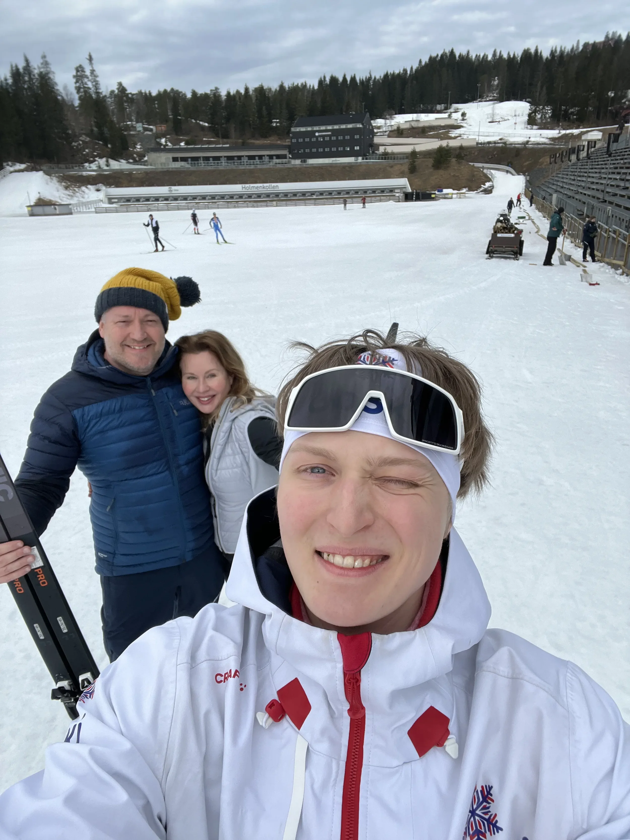 Three people take a selfie on snow skis on a ski slope. Two of them smile at the camera, one further back appears to be putting on ski gear. In the background are several people skiing on the snow, as well as buildings and trees in a mountain setting.